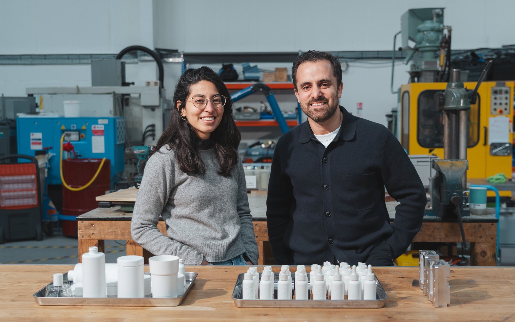 A smiling portrait of Shellworks co-founders, Insiya and Amir, standing behind a workbench in their functional innovation workshop. In the foreground, metal trays display pristine white VIVOMER prototypes, including bottles, tubs, and droppers, sitting alongside complex multi-part metal injection moulding tools. This image highlights their hands-on leadership and vertical expertise in translating biotech innovation into retail-ready, scalable sustainable manufacturing solutions.