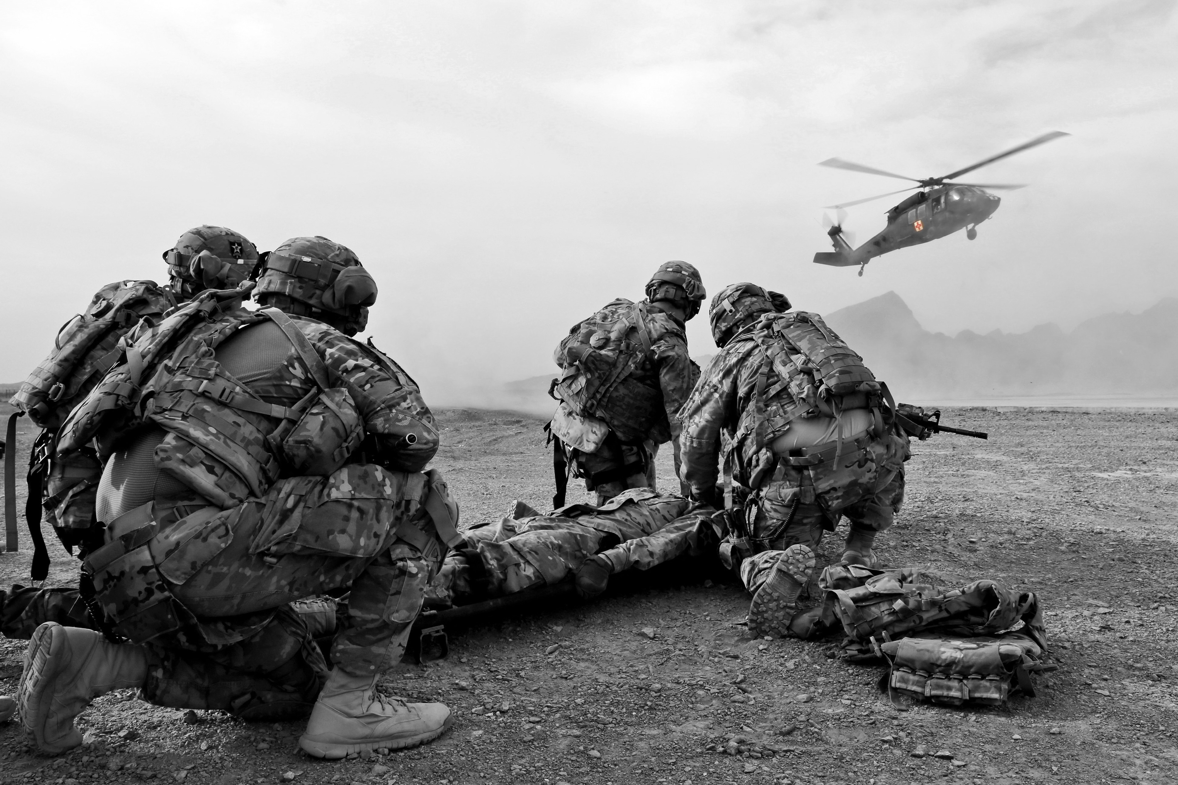 Black and white photo of 4 military paramedics kneeling near a wounded soldier. On the background a medical evacuation helicopter is approaching.