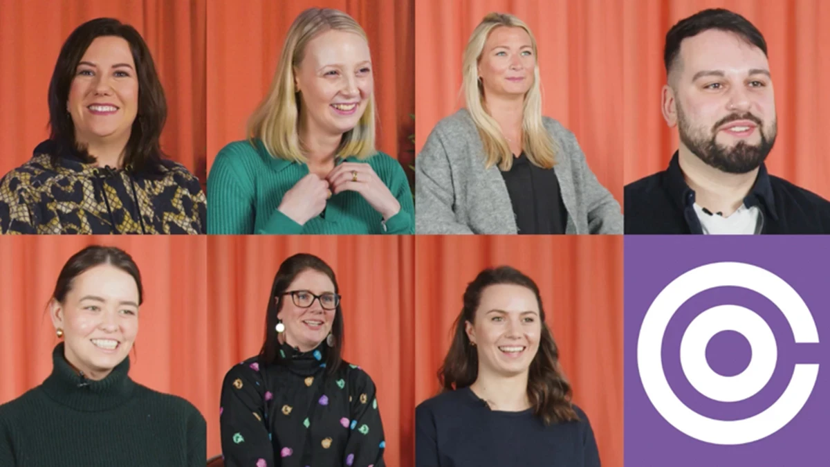Group of eight individuals arranged in two rows in front of an orange curtain, with a purple circular logo in the bottom right corner.