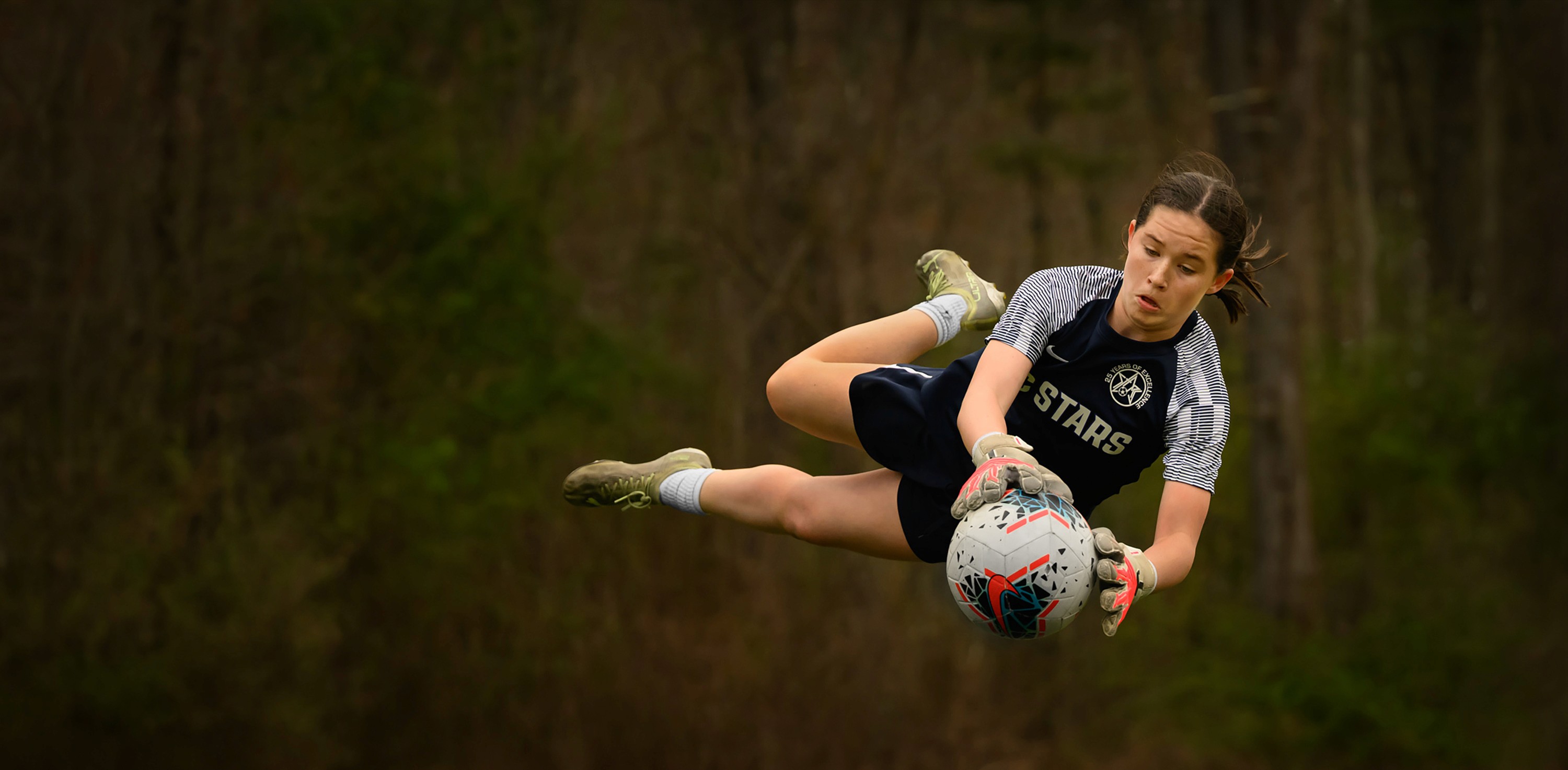 A female youth goalkeeper leaps through the air holding the ball against a dark background, showcasing agility and athleticism.