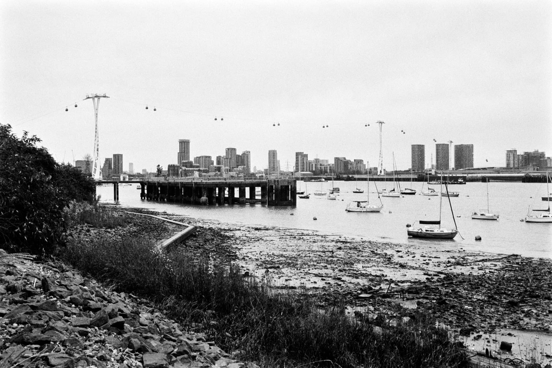 Thames foreshore with moored sailboats at low tide, wooden pier structure extending into river, Emirates cable car crossing overhead, Canary Wharf development skyline beyond, rubble and vegetation in foreground