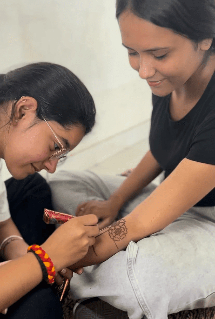 Female college student smiling while having intricate mehandi applied to her forearm by a peer.