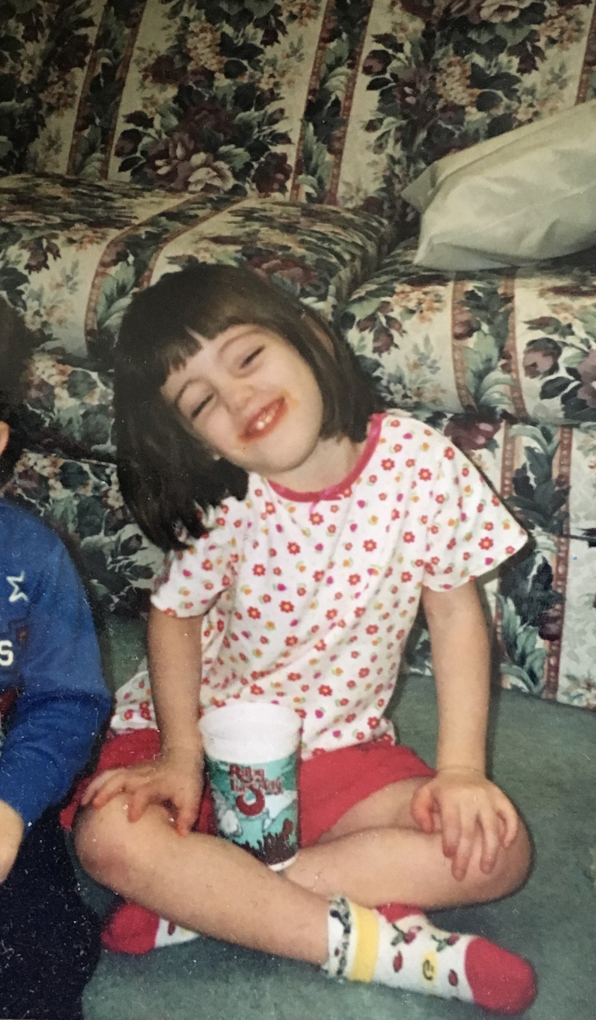 Childhood photo of Emily with silly smile, squinted eyes, and head tilted to the left seated on a floor in front of a couch