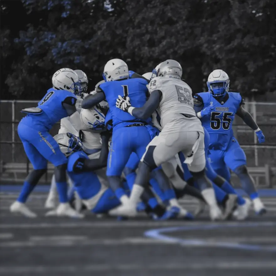 A group of football players in blue uniforms tackles a player in gray on the field.