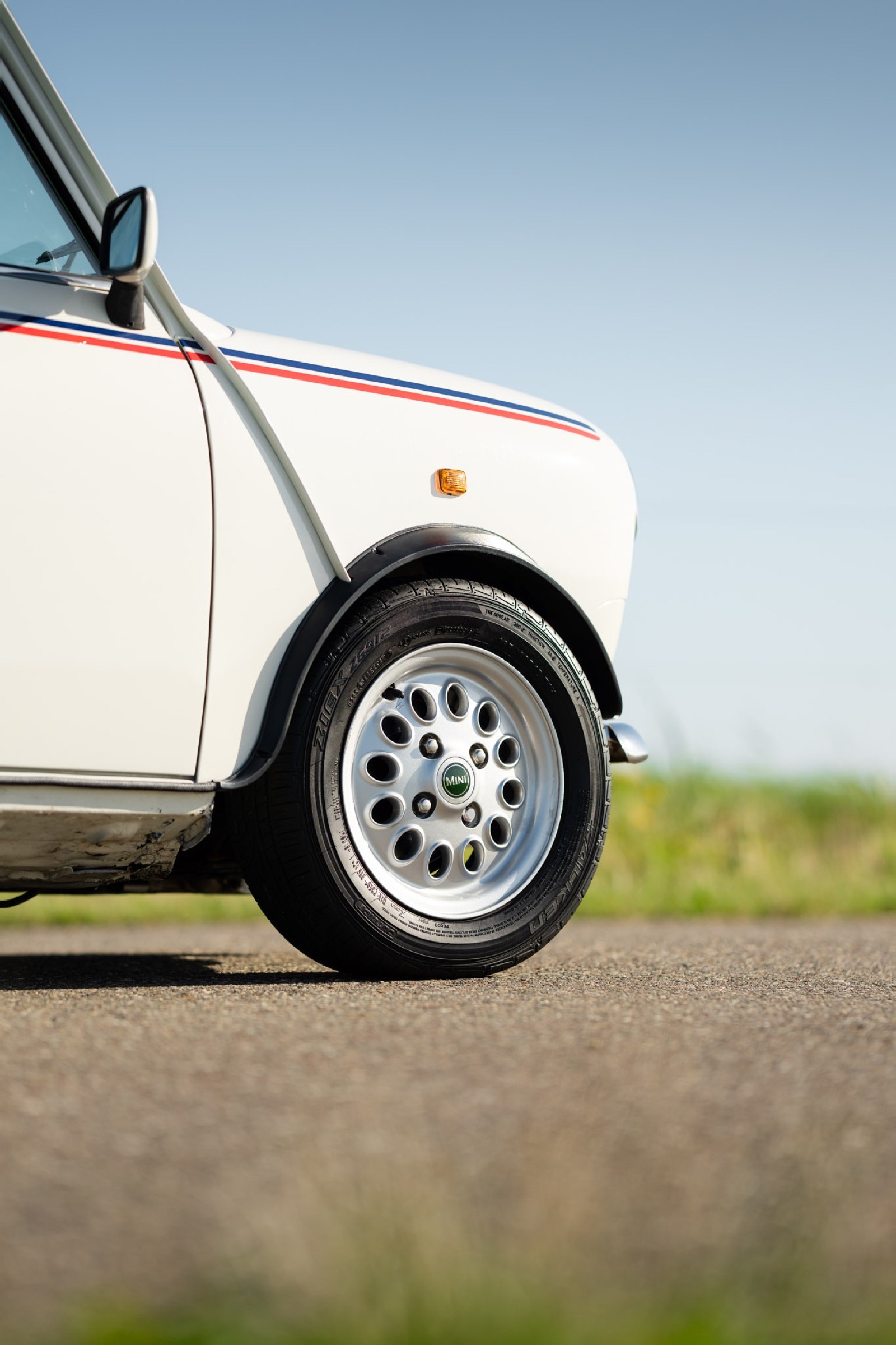 Close-up of a car's white wheel and tire, with a blurred grassy background and clear blue sky above.