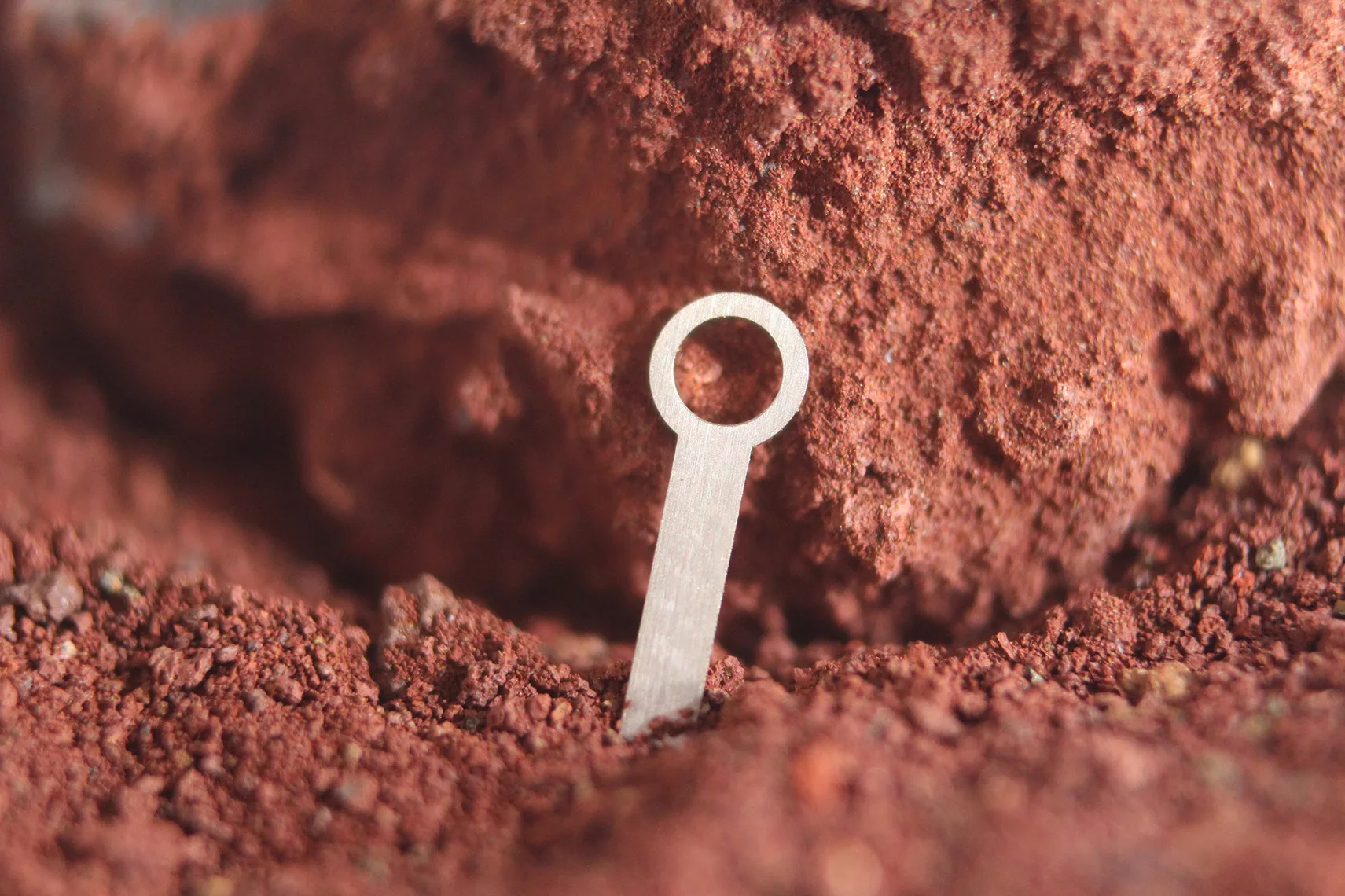 High-resolution macro photograph of a tiny, silver metal component (possibly a watch hand or indicator) standing upright in rich, red-brown mineral powder or dirt. Focus on industrial precision, material contrast, and micro-scale engineering.