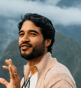 A smiling person with curly hair sits at a desk, looking at a laptop against a dark background.