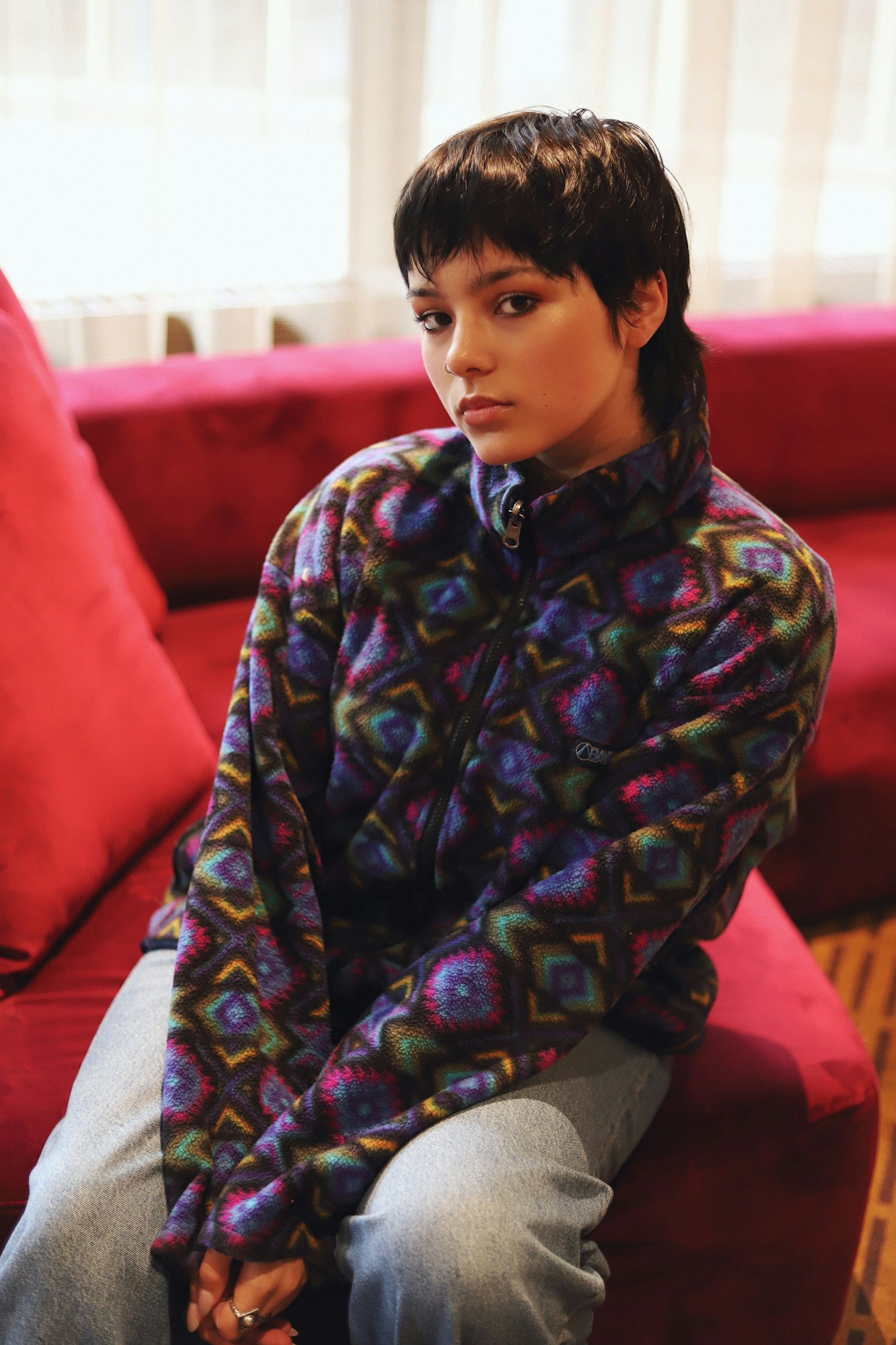 A portrait of the person sitting on a red velvet sofa, looking directly at the camera with a neutral expression. They are wearing the same patterned fleece jacket and light-wash jeans.