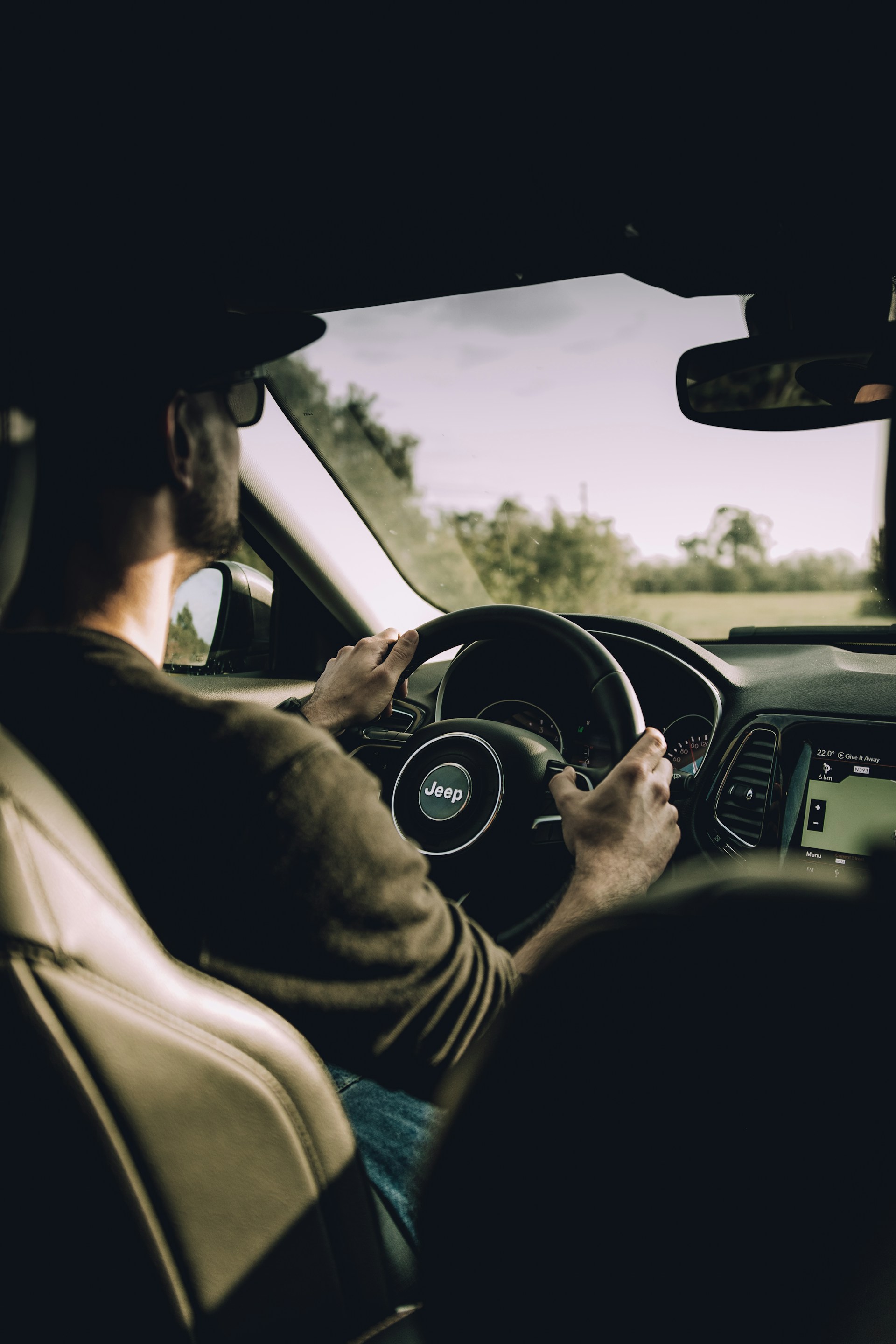  An interior shot from behind the driver, showing a man wearing sunglasses and a baseball cap driving a Jeep. His hands are firmly gripping the wheel. The car is on a road surrounded by green trees and fields visible through the windshield, under bright natural light.