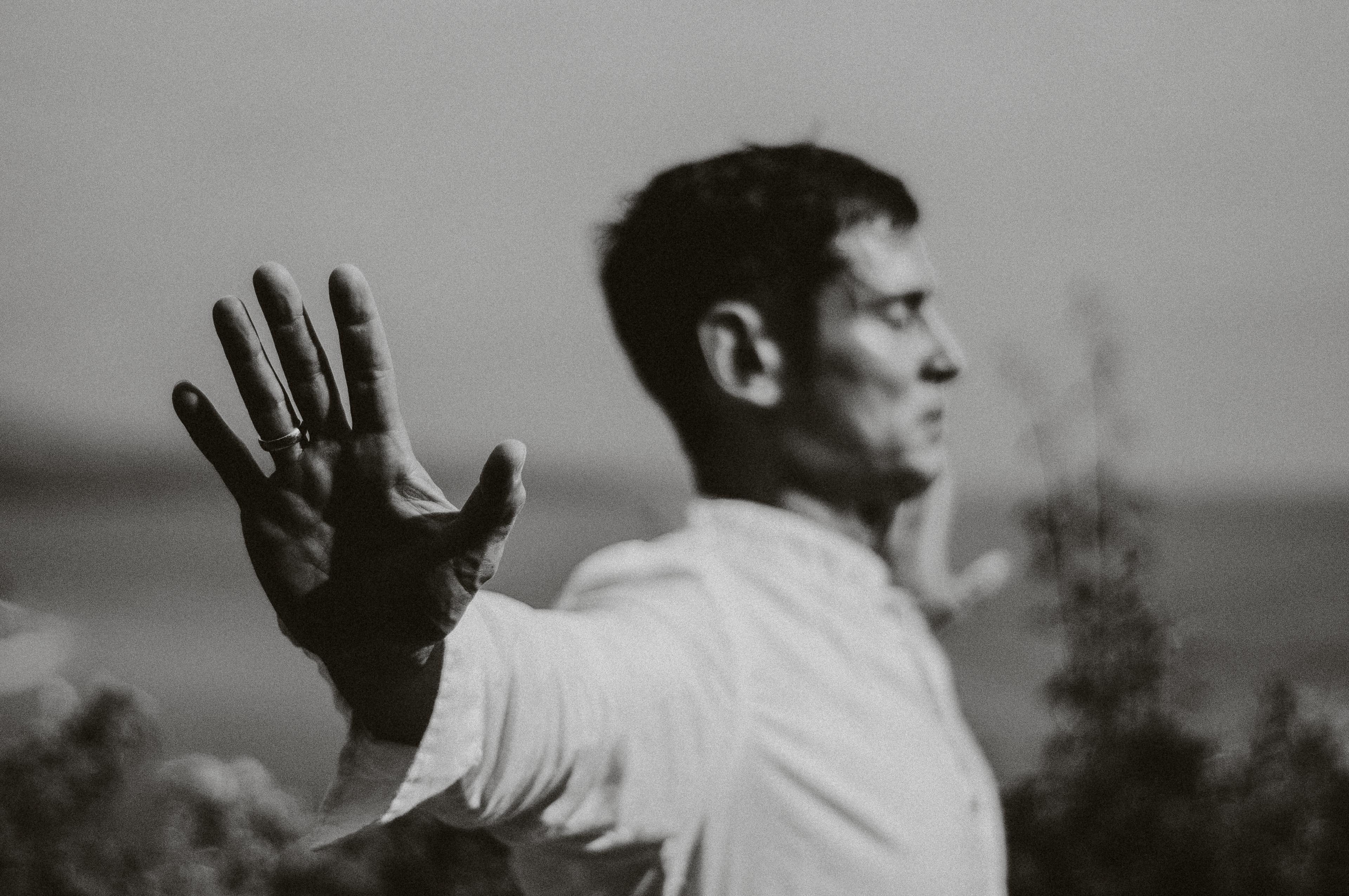 A black and white photo of a person standing outdoors in a serene setting, with their hand extended towards the camera, wearing a white shirt, embodying a calming and reflective pose.