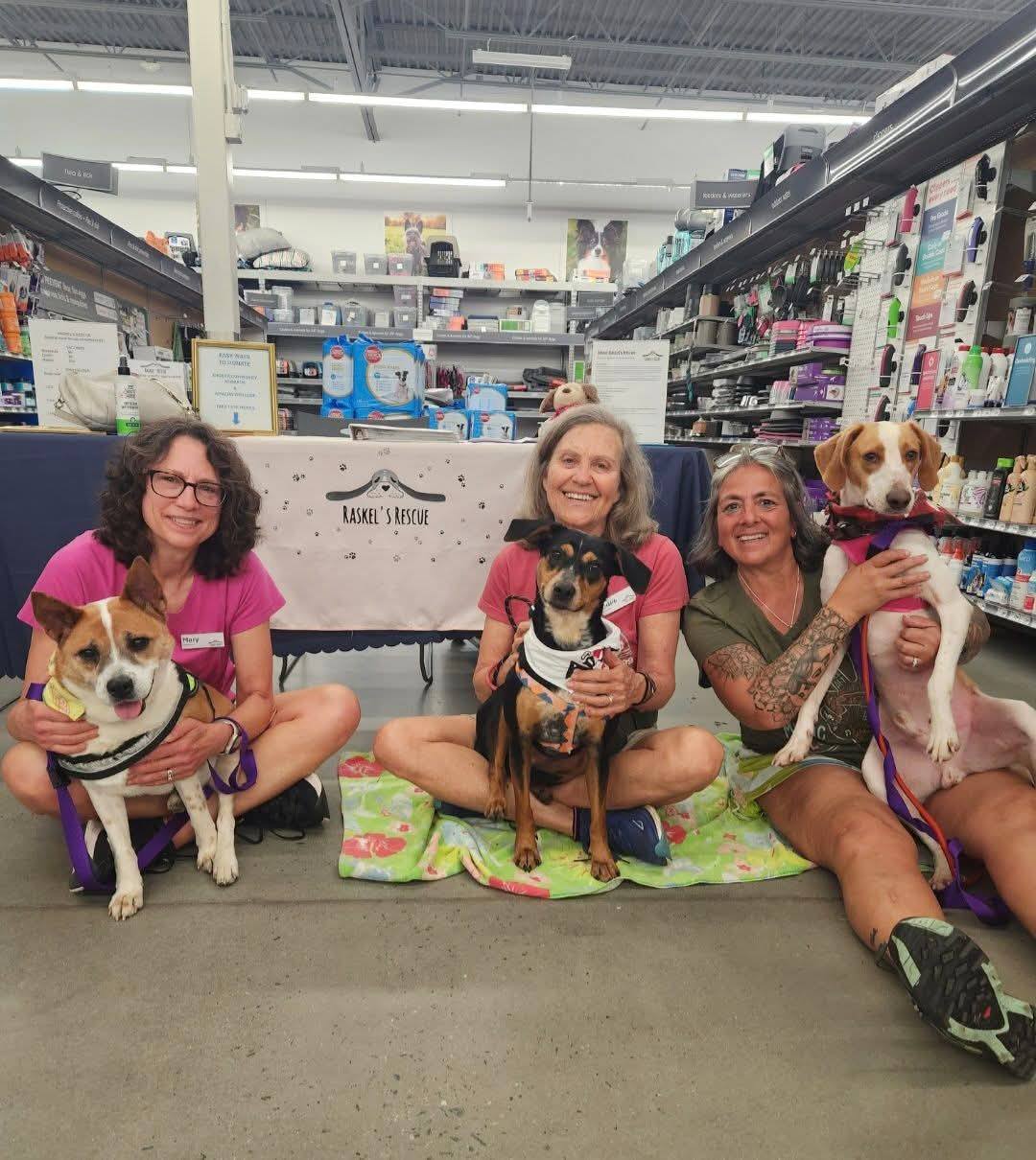 Four women sit on the floor in a store, each holding a dog, surrounded by pet supplies and smiling happily.