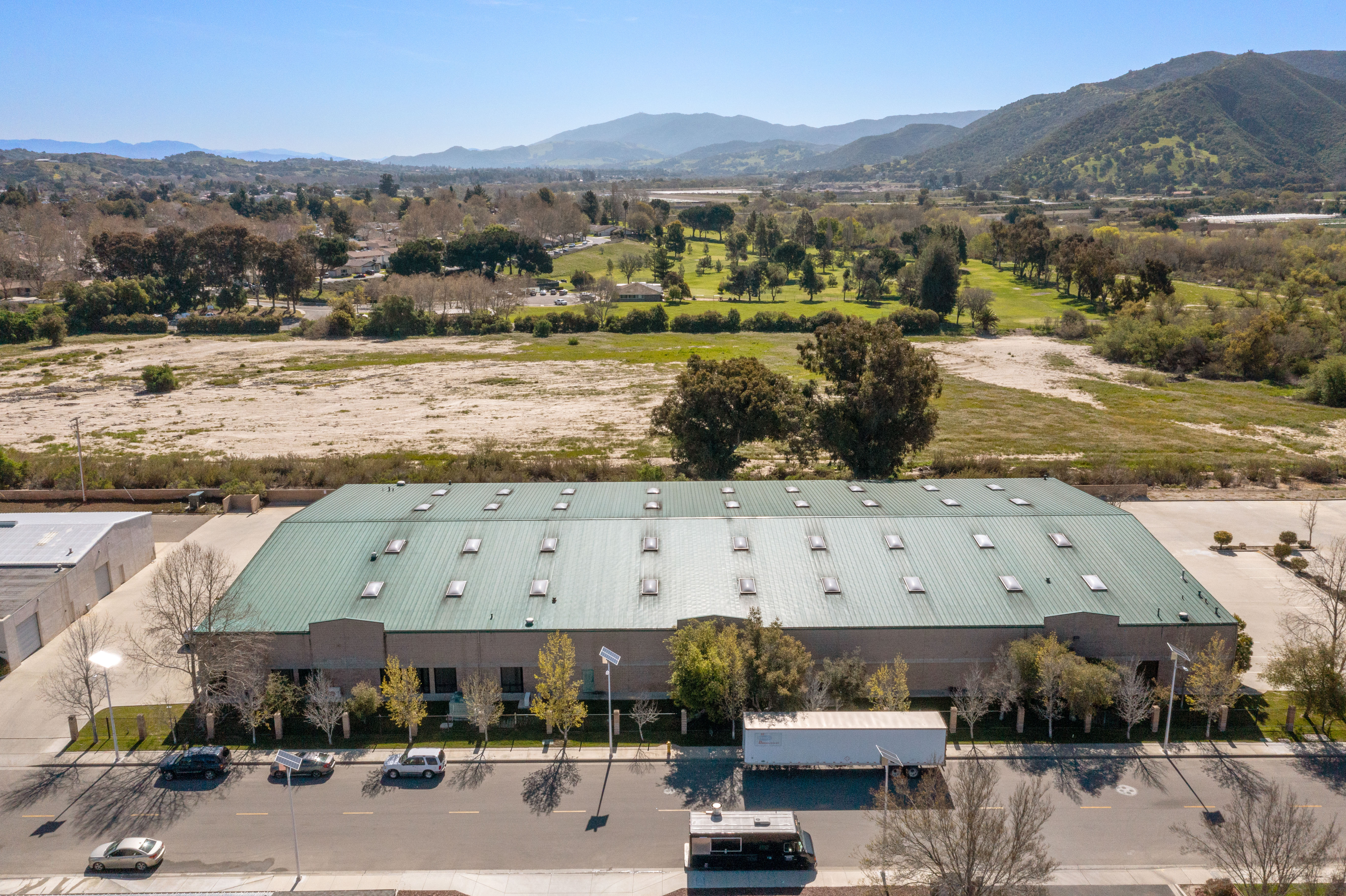 Aerial view looking toward the front facade of the warehouse building, with a green metal roof, front parking lot with mature trees, and an open green space and golf course visible behind the property against a mountain backdrop.