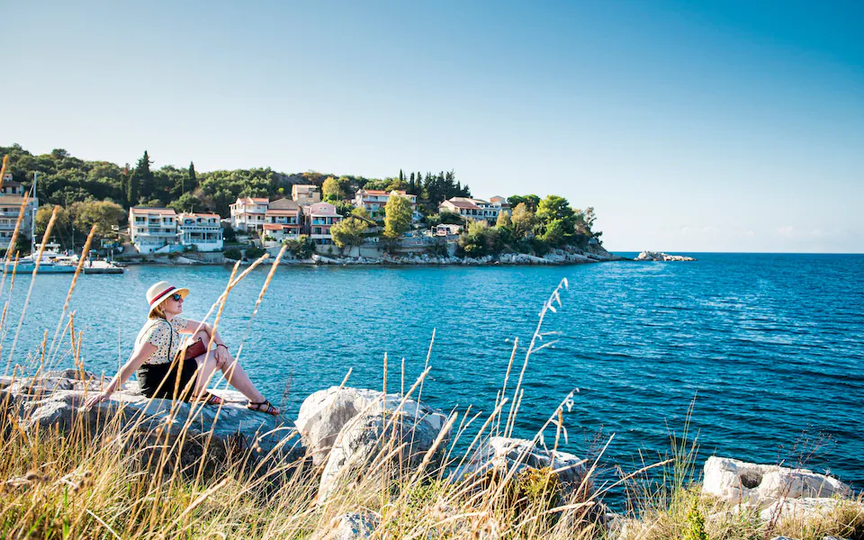 A woman wearing a sunhat and sunglasses sits on large rocks by the sea, looking out over a calm blue bay. 