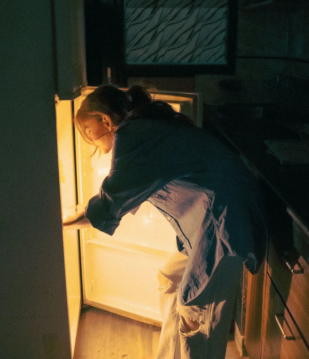 Woman looking inside an open refrigerator at night in a dimly lit kitchen.