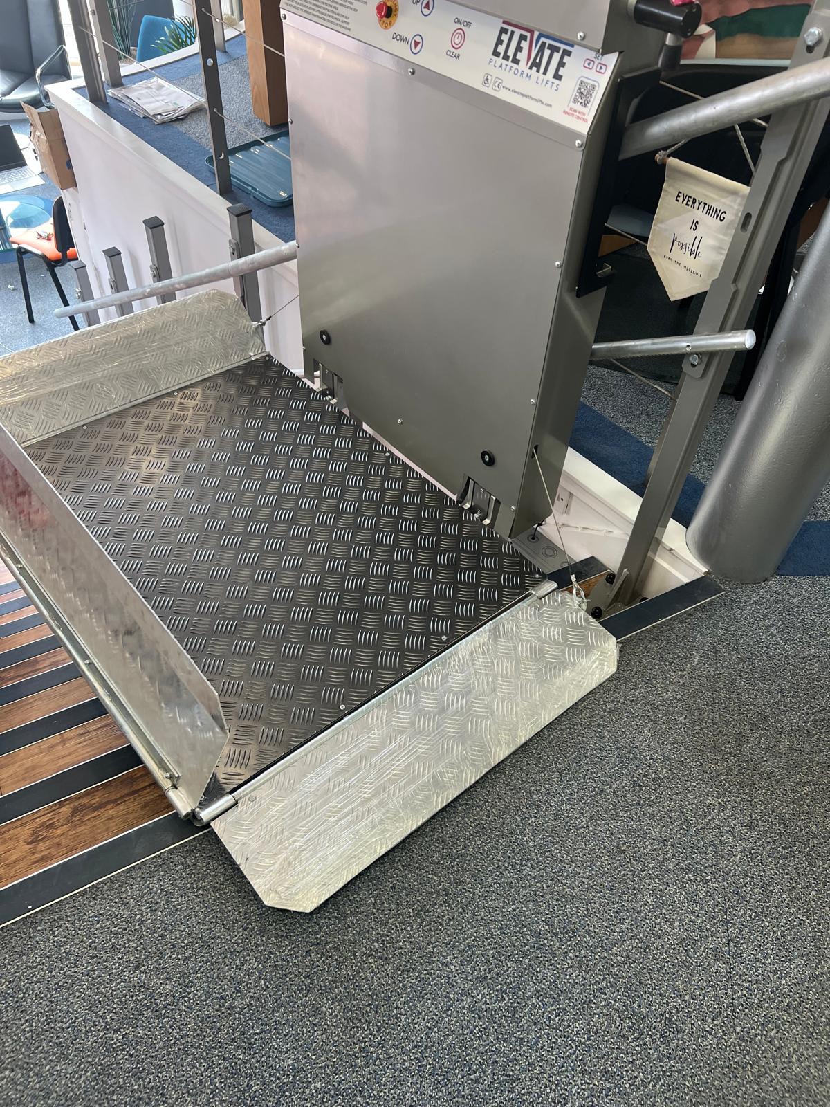 Elevate Platform Lifts vertical step lift at lower landing position in a showroom setting — large anti-slip chequer plate platform with fold-out access ramp, control panel with up/down buttons, and staircase handrail visible alongside