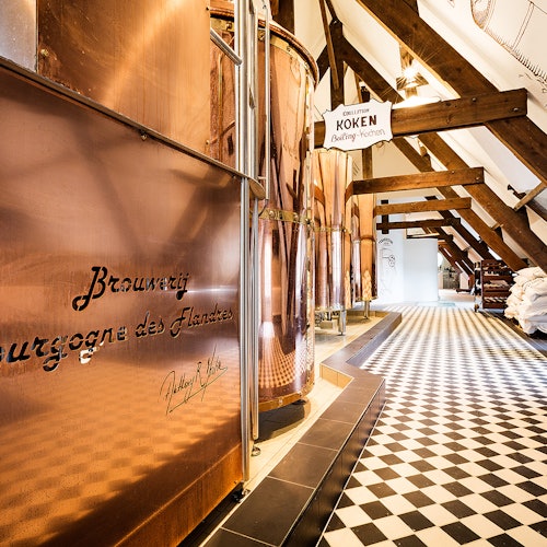 Et bryggeriets interiør med store kobbertanker, svart-hvitt rutete gulv, og et skilt med teksten "KOEKEN" over tankene.