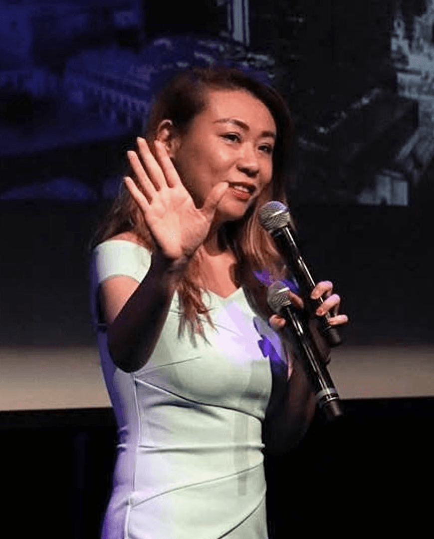 oman speaking on stage with a microphone, smiling and raising her hand.