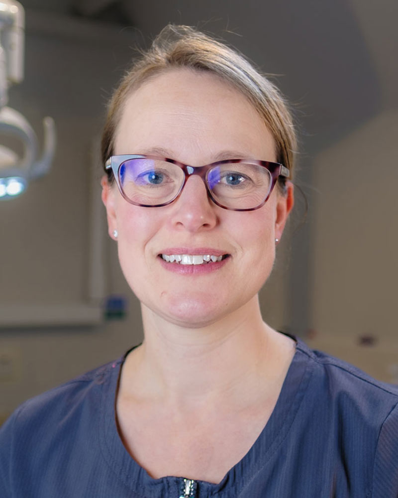 A portrait of Emma, Associate Dentist at Cricklade Dental Practice, smiling and wearing glasses and a dark grey scrub top with yellow embroidered text that reads "Emma" on the left and "Cricklade Dental Practice" on the right. She is standing in a dental surgery with a dental light visible in the background.