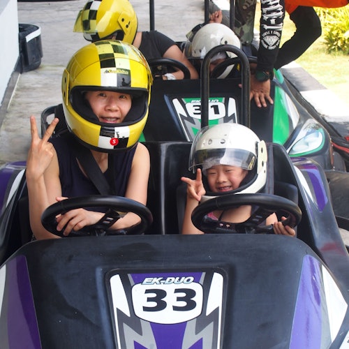 Two people in helmets sitting in go-kart number 33, both making peace signs. Others are visible in the background.
