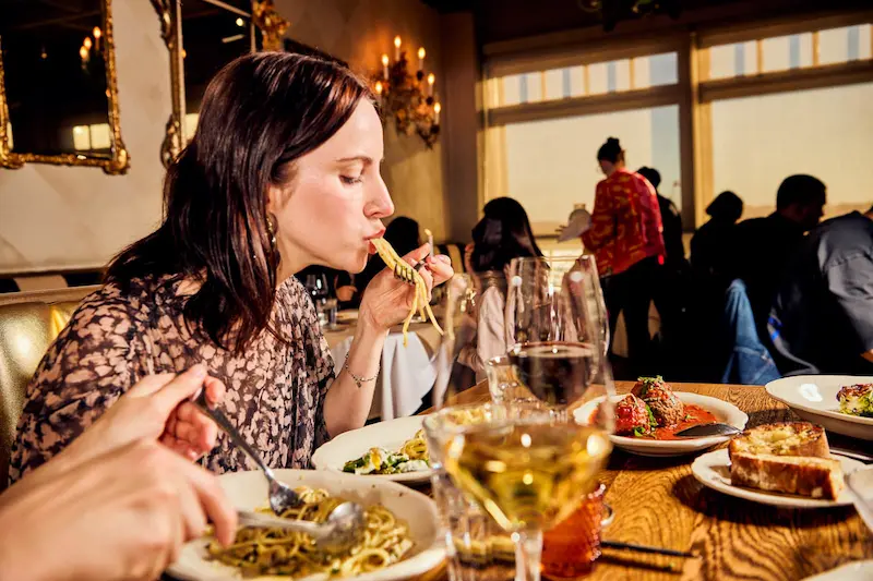 A woman eating pasta at a busy restaurant table with wine and appetizers.