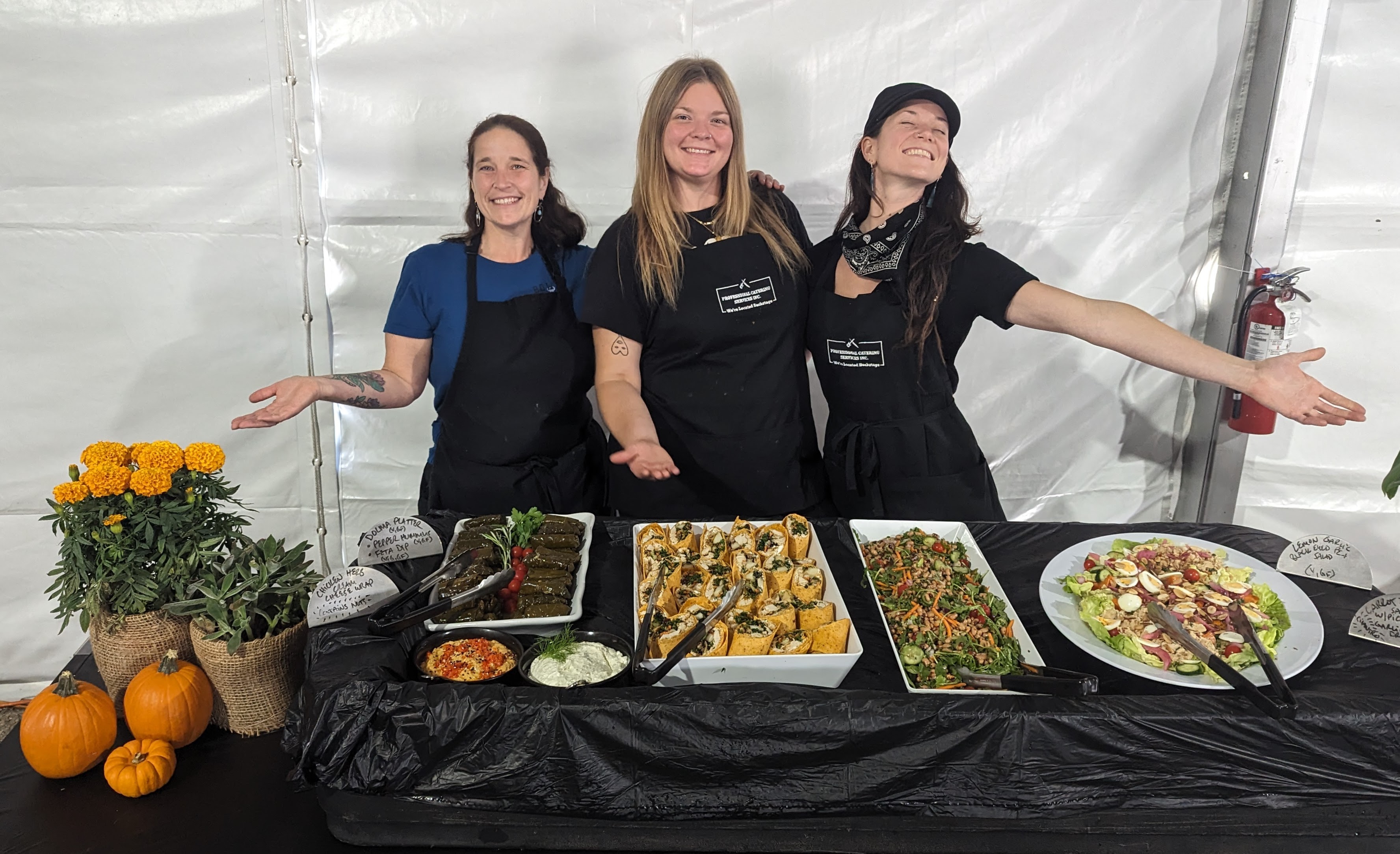 Three women stand behind a table filled with various dishes, smiling and gesturing happily at a food event.