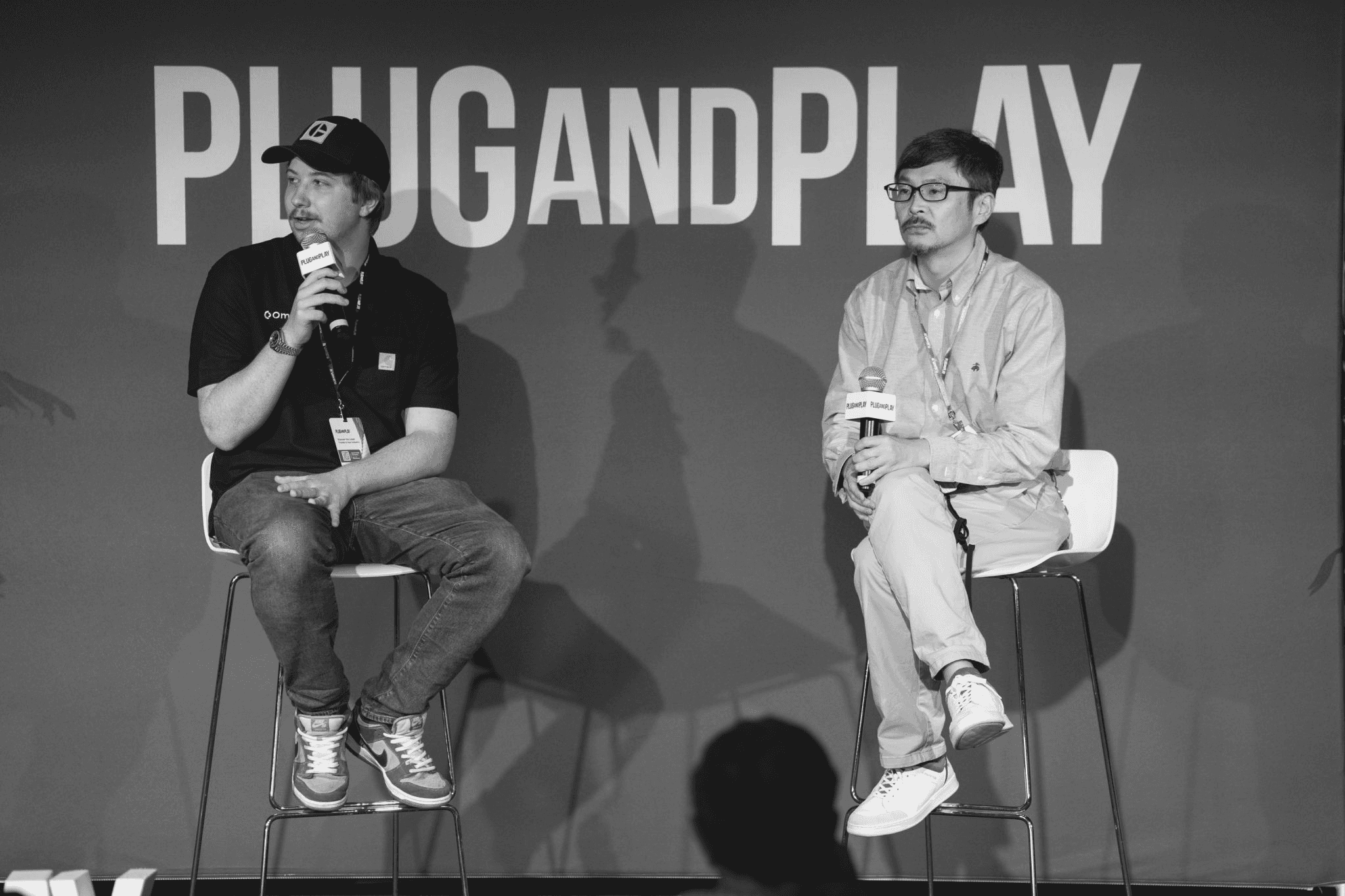 Photo of two men sitting on chairs at the "Plug and Play" conference with microphones speaking to an audience