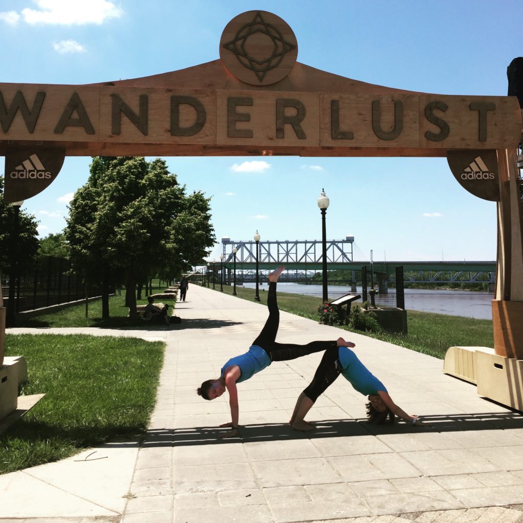 Two people perform a yoga pose under a wooden archway labeled "Wanderlust," set against a backdrop of a sunny riverside park with a clear blue sky.