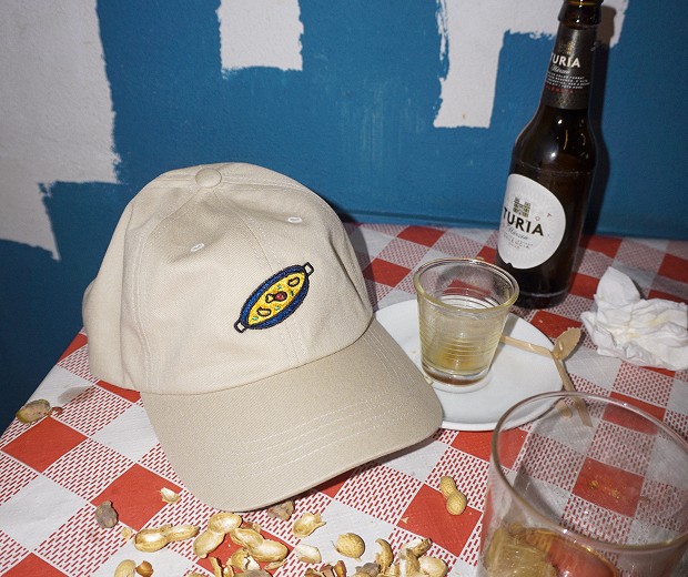 Cap with an embroidered paella patch on a checkered tablecloth next to a Turia beer.