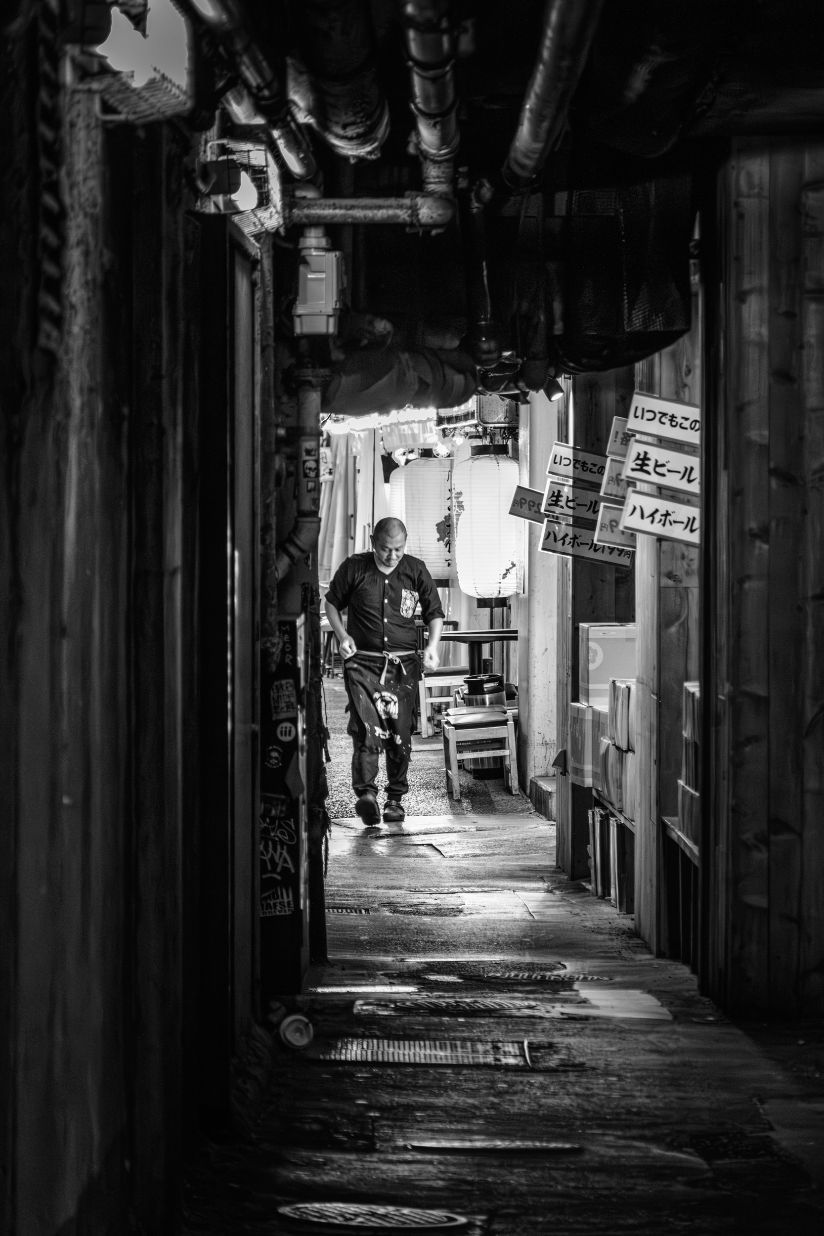 Black and white photograph of a cook walking inside a narrow alley lined with pipes, signs, and lanterns.