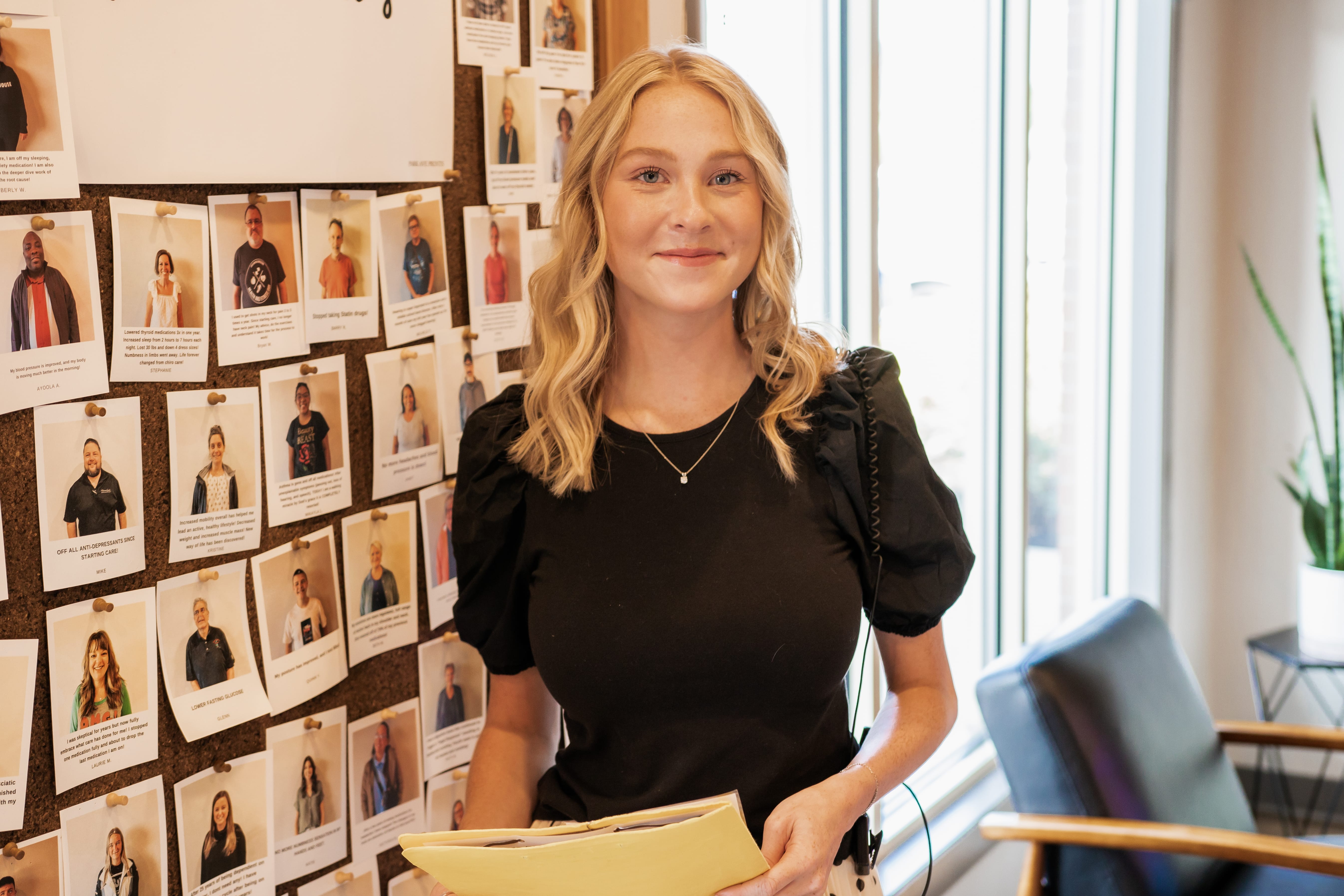 A female smiling Premier employee in an office standing next to a wall of photos of Premier clients.