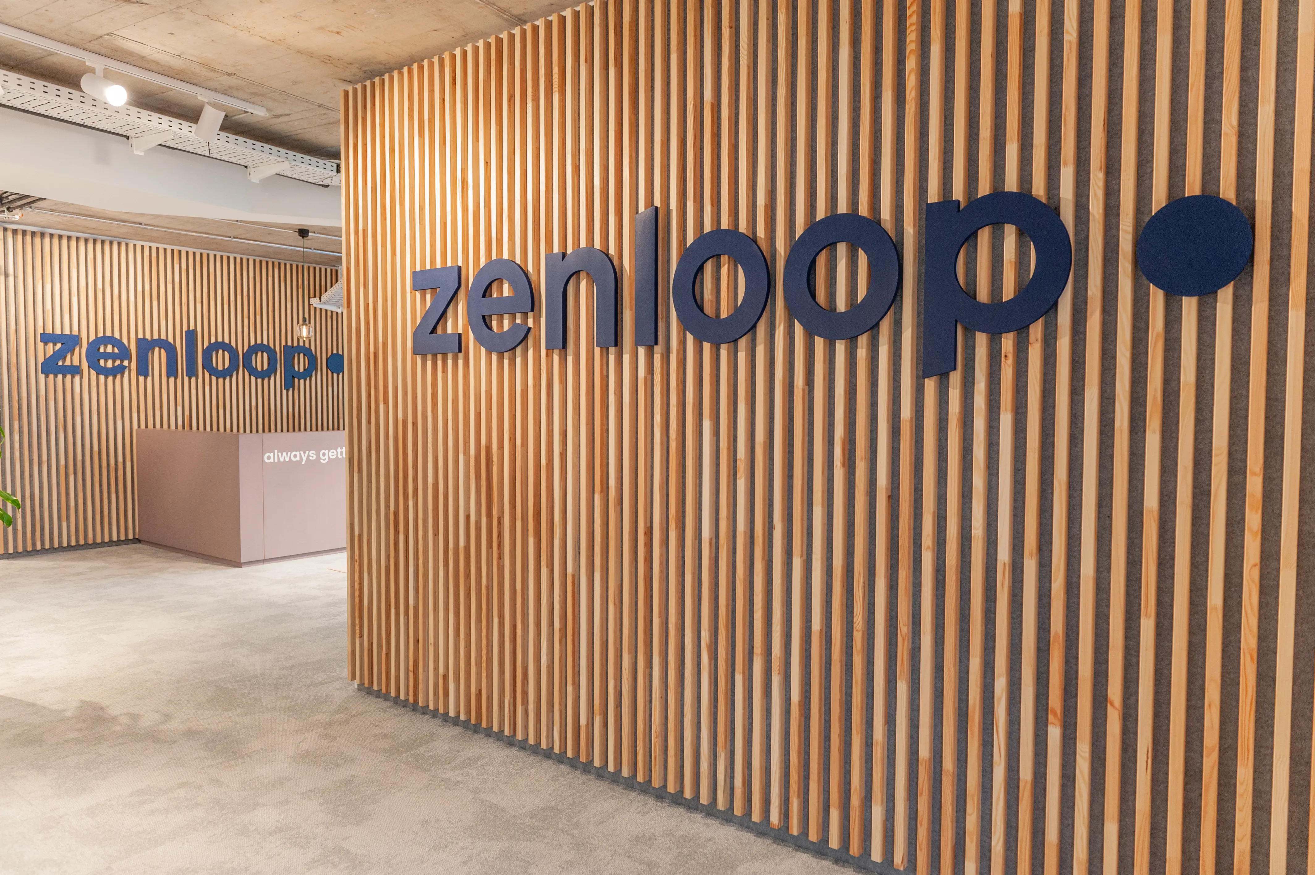 Entrance at ZENLOOP with a navy 3D logo mounted on a floor-to-ceiling wooden slat wall, a second logo above the dusty-pink reception desk and polished concrete flooring.