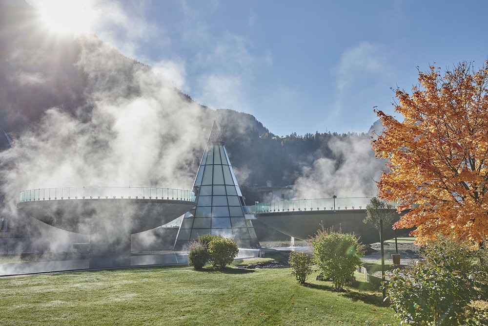 Eine Frau im weißen Bademantel steht neben einem dampfenden Außenpool im Aqua Dome Therme Längenfeld, umgeben von schneebedeckten Bergen und nebligem Himmel.