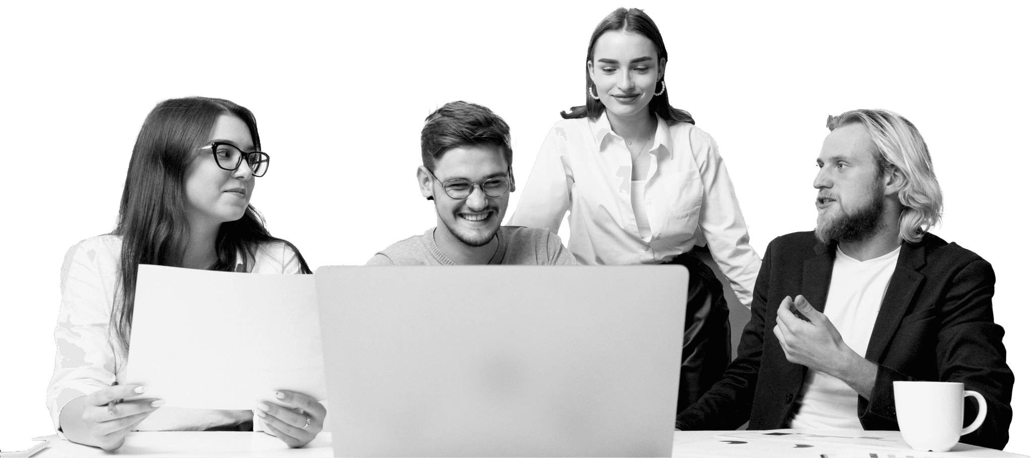 A group of four people are gathered around a laptop at a desk, collaborating and discussing work, each with engaged expressions, with papers and a coffee cup visible.