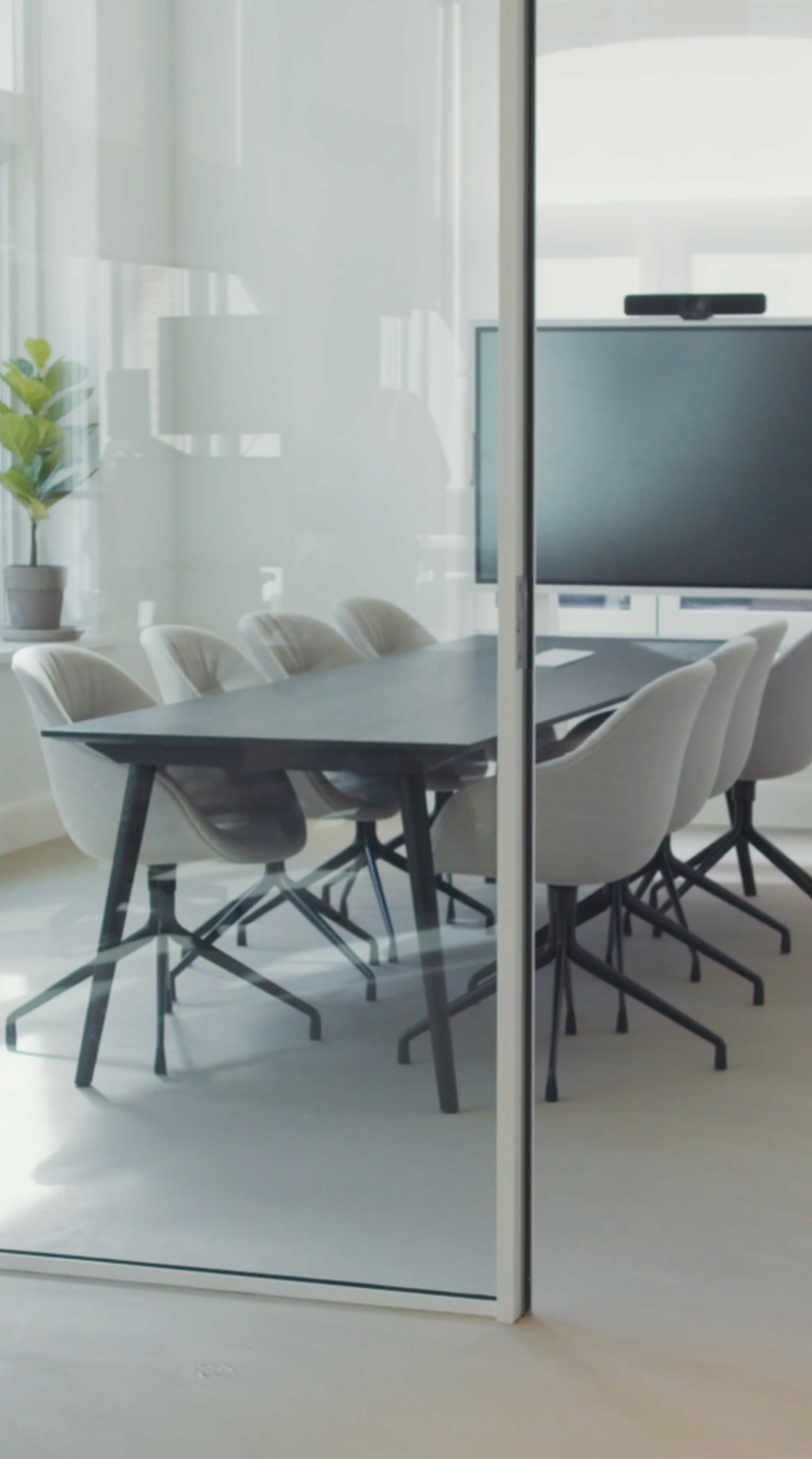 Modern meeting room with a round table, gray chairs, and a large monitor, viewed through glass.