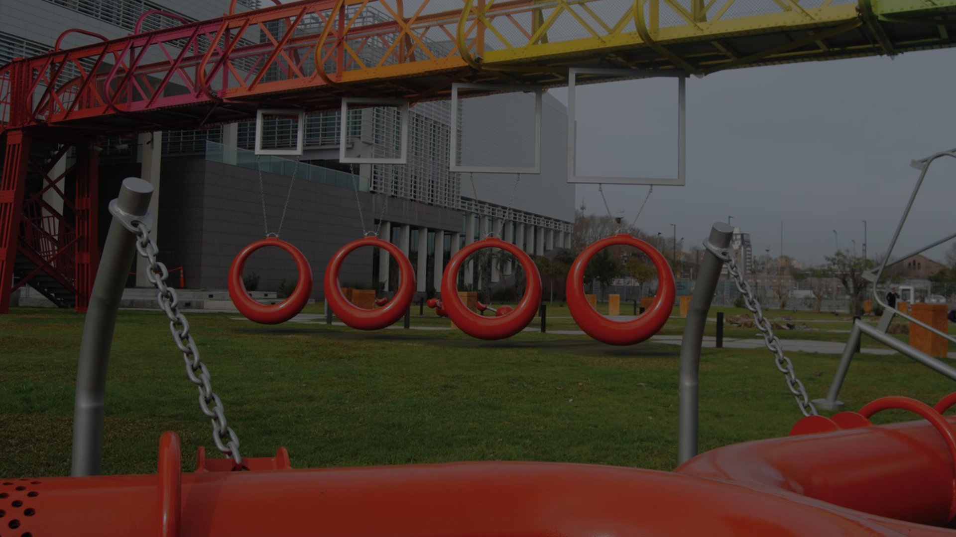 Red circular playground swings hang below a vibrant, rainbow-colored bridge structure, set in a grassy park beside a modern building, suggesting playfulness.