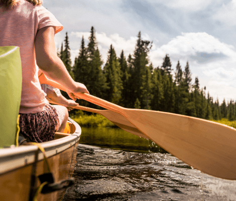 A close-up view of a person kayaking on a calm river, with a wooden paddle in hand. The individual is wearing a pink shirt and sitting in a kayak, with lush green trees lining the shore in the background. The sky is partly cloudy, creating a serene outdoor atmosphere.