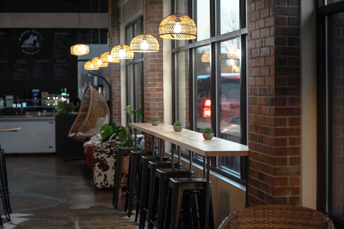 A thick cut of steak with prominent grill marks sizzles over hot coals and flames on a barbecue, with blurred figures of people and drinks in the background, suggesting an outdoor gathering.