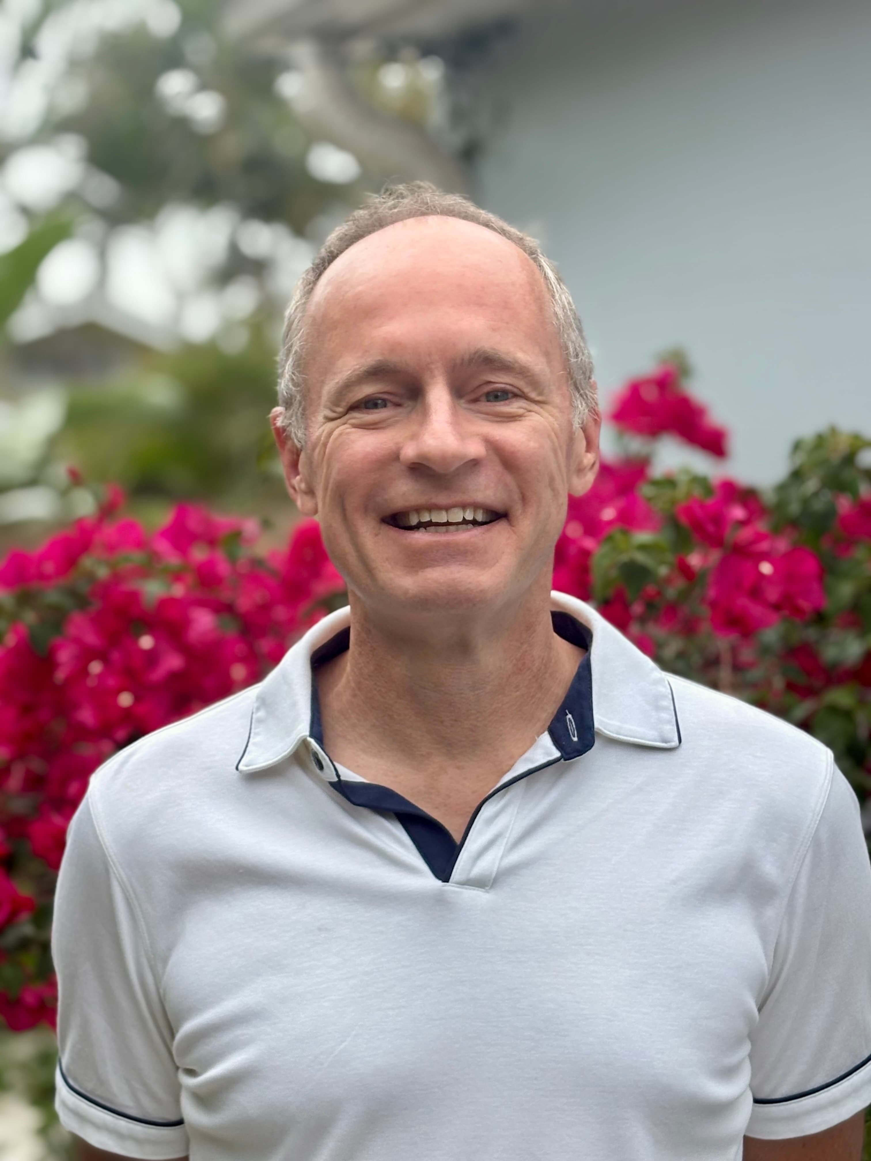A smiling man with glasses, curly hair, wearing a maroon hoodie, poses against a dark background.