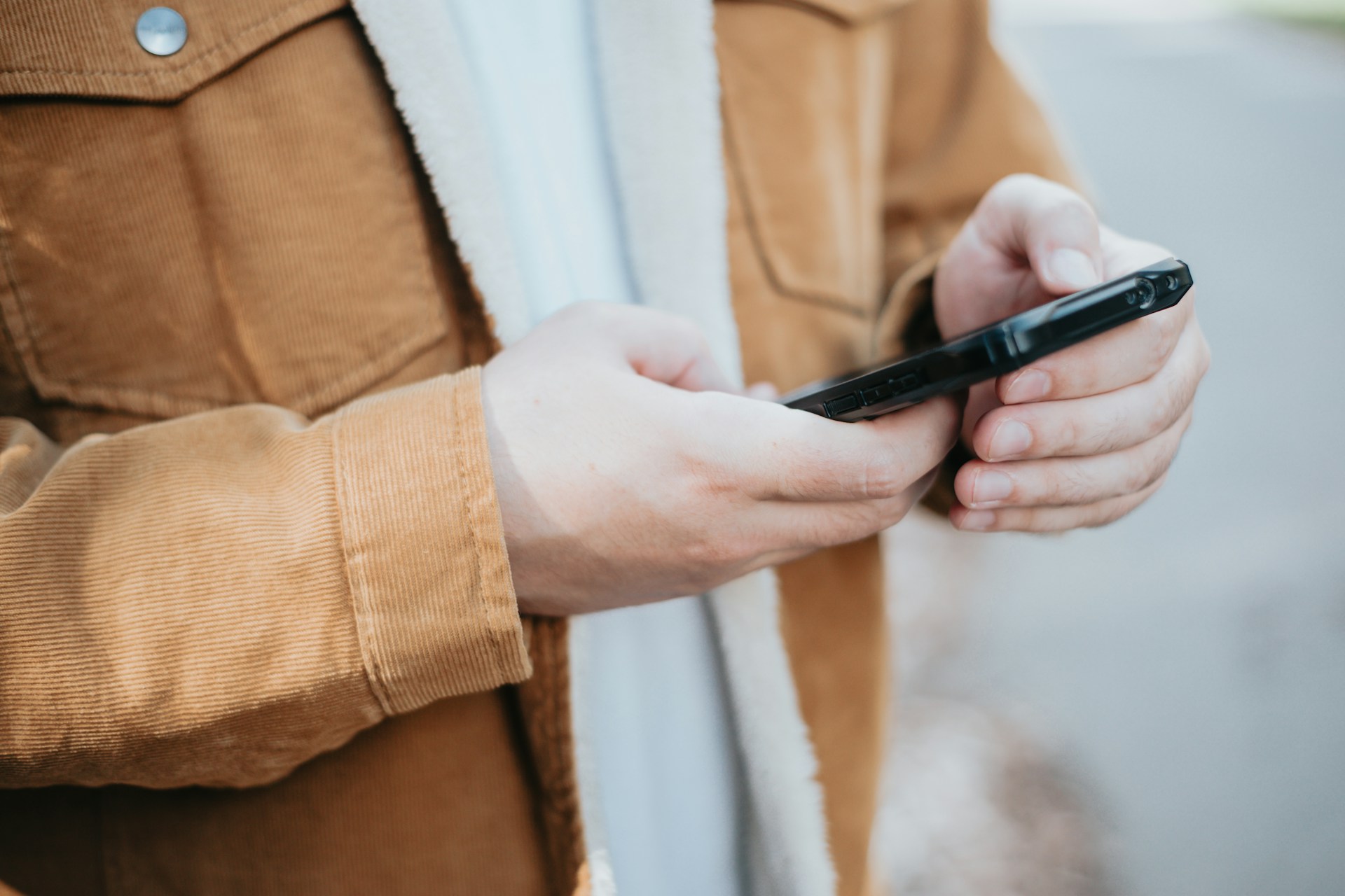 Close-up of a person wearing a tan jacket and holding a smartphone outdoors, focused on their hands and device.