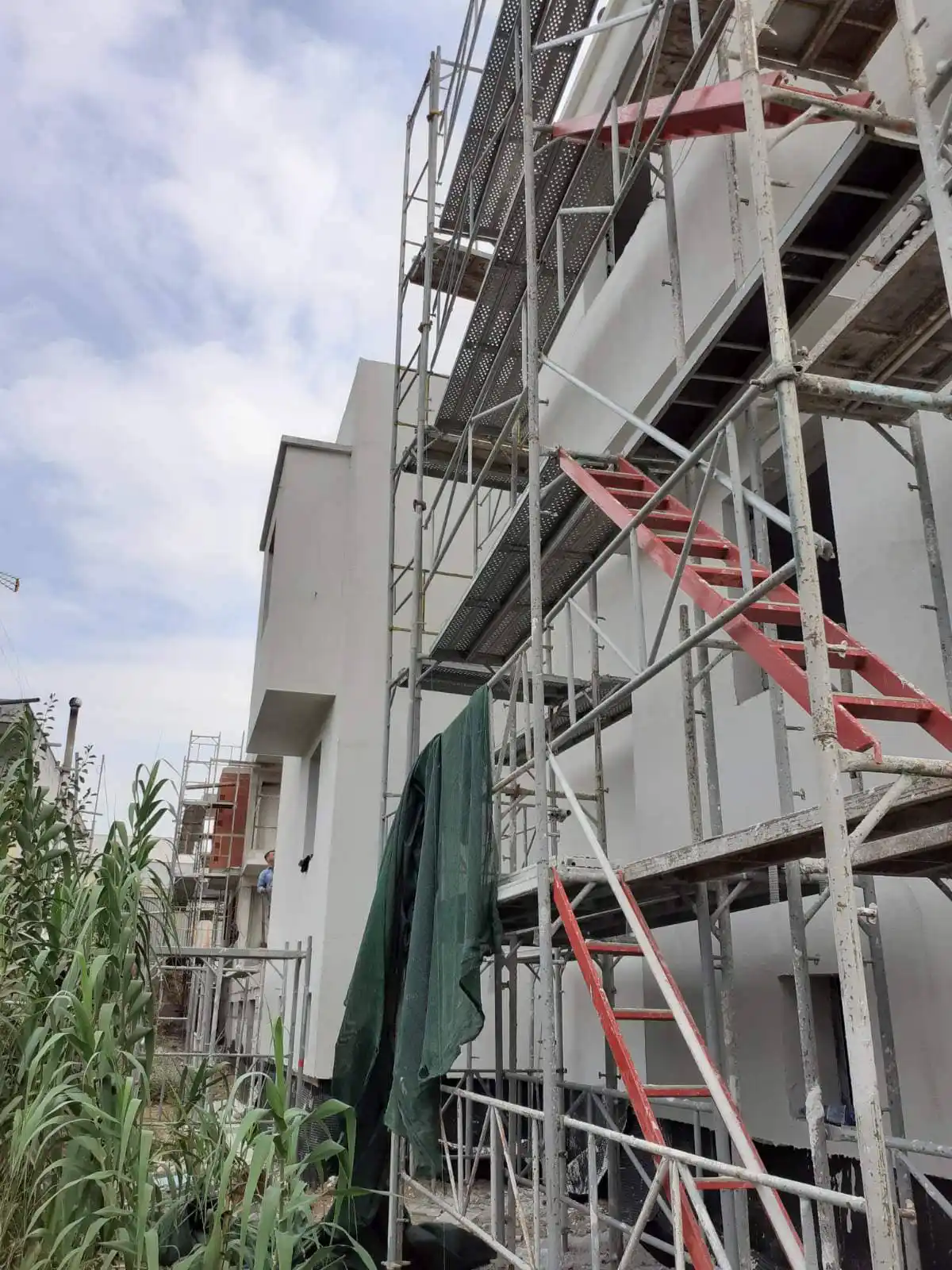 Εξωτερικές μεταλλικές σκαλωσιές με κόκκινες σκάλες σε πρόσοψη λευκού κτιρίου υπό κατασκευή.