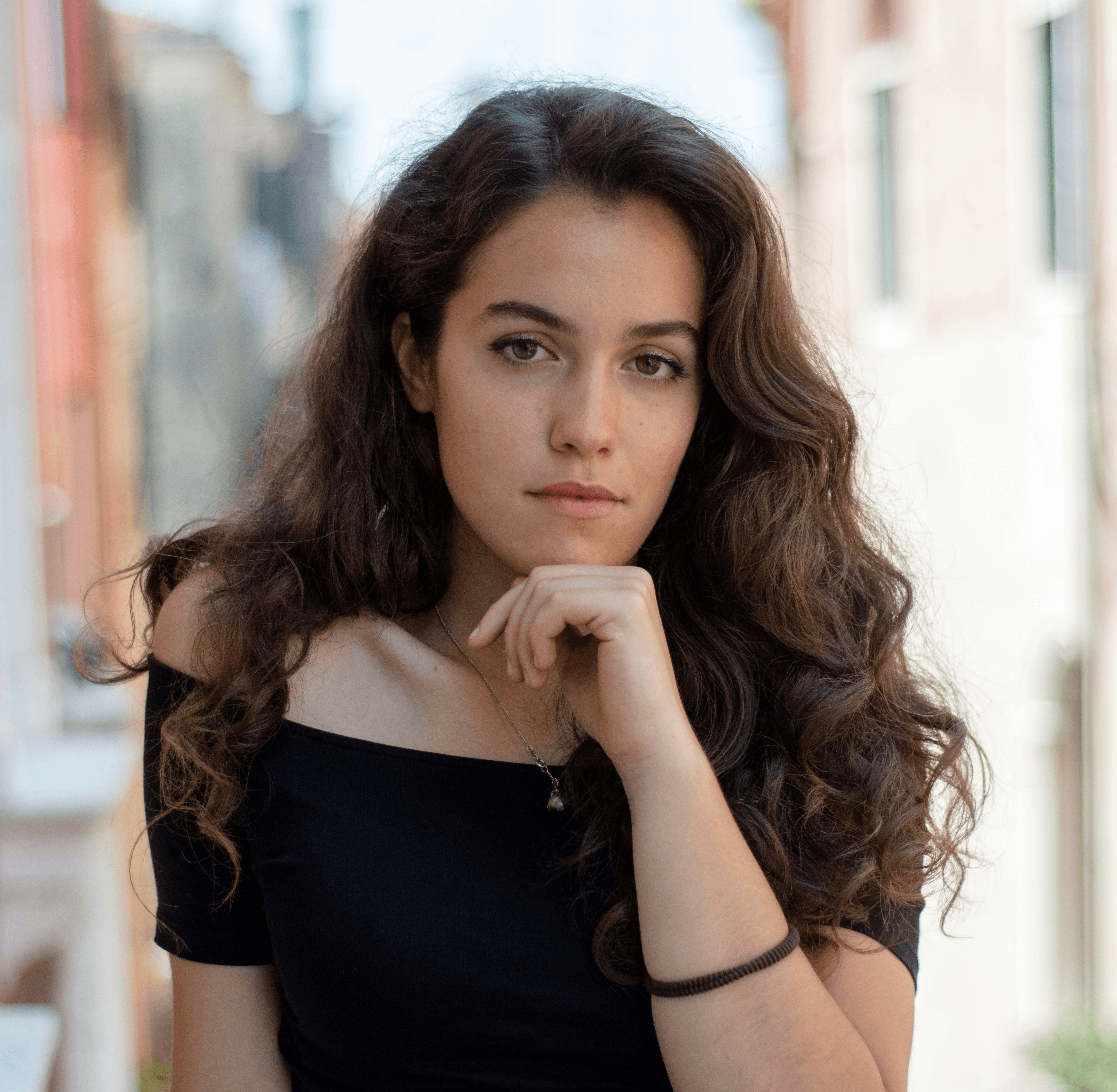 Person with long curly brown hair in black off-shoulder top posing outdoors