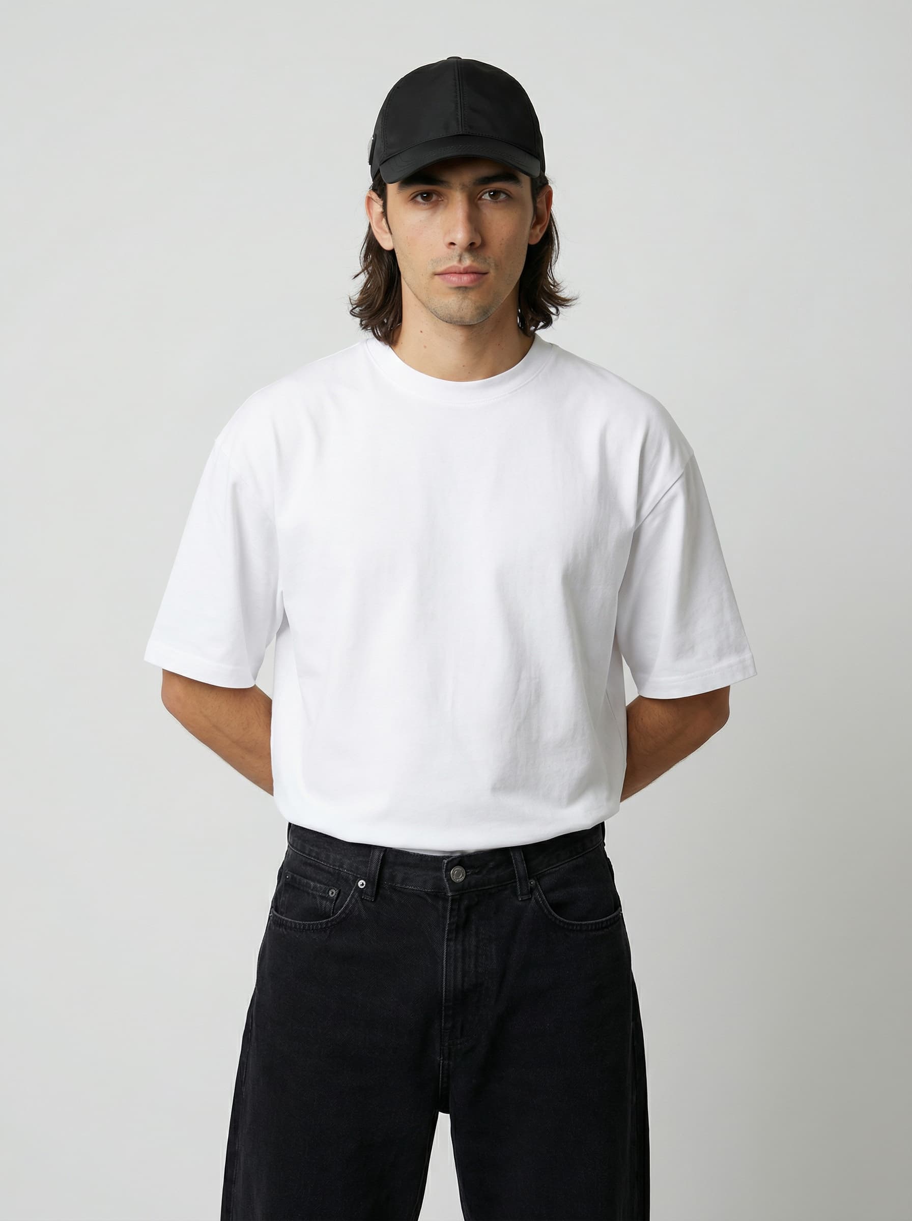 A person wearing a plain white t-shirt, black jeans, and a black cap stands against a neutral background.