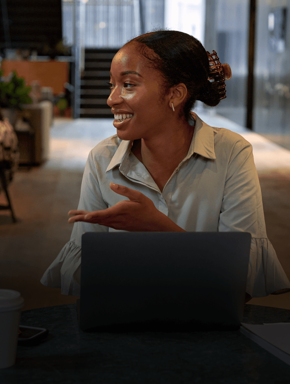 Photograph of a woman sitting at a table at work with her laptop in front of her, turned to the side and talking to someone