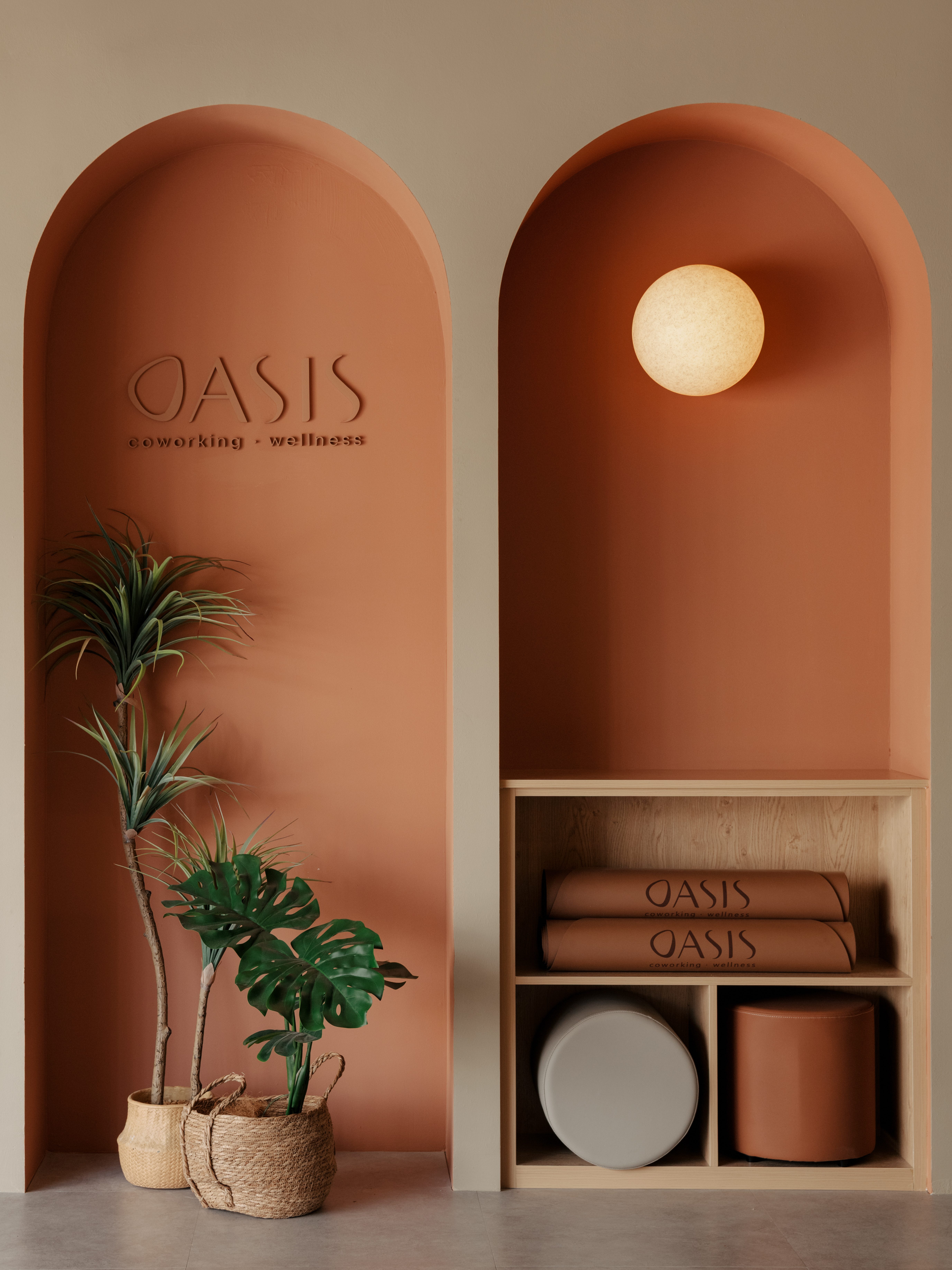 A cozy interior of the OASIS coworking space featuring terracotta arches, a modern light fixture, and plants, with a shelf displaying wellness items and decorative elements.