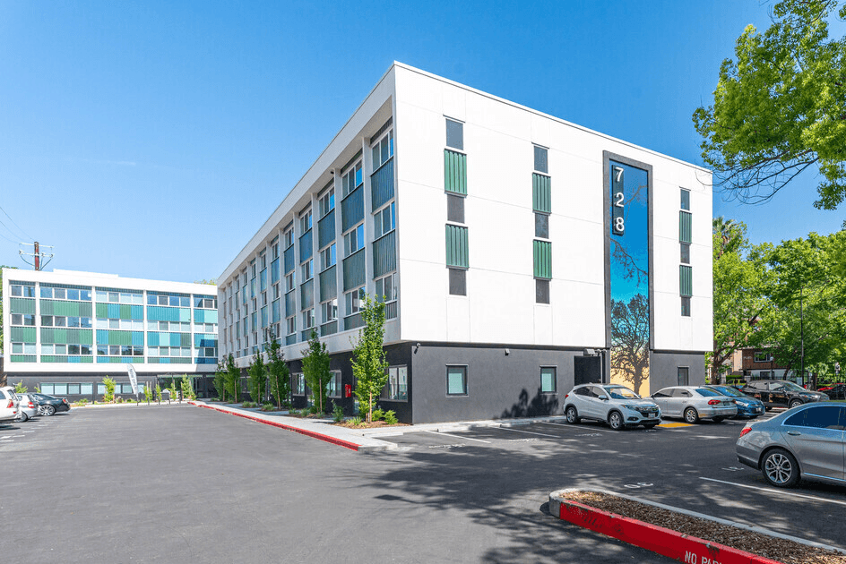 A four-story apartment building in Midtown Sacramento features a large mural with a tree and the number "728" displayed vertically. The black and white exterior walls house modern living spaces, boasting numerous sun-drenched windows. Two cars are parked in the lot, surrounded by trees.