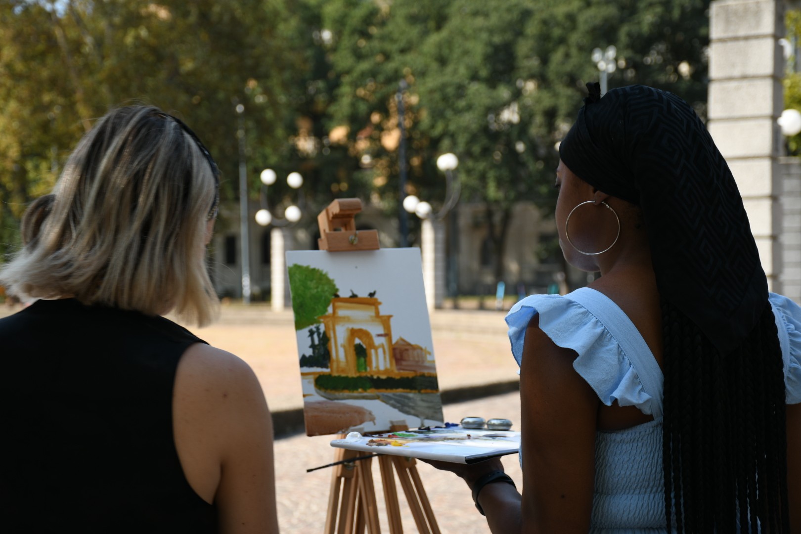 Two individuals stand outdoors, engaged in painting a scenic landscape on a canvas, with a pathway and surrounding trees, suggesting an art session in a park setting.