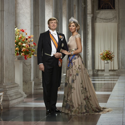 En man och en kvinna i formell klädsel poserar i en utsmyckad sal med blommor och kolonner. Kvinnan bär en tiara och en blommig klänning.