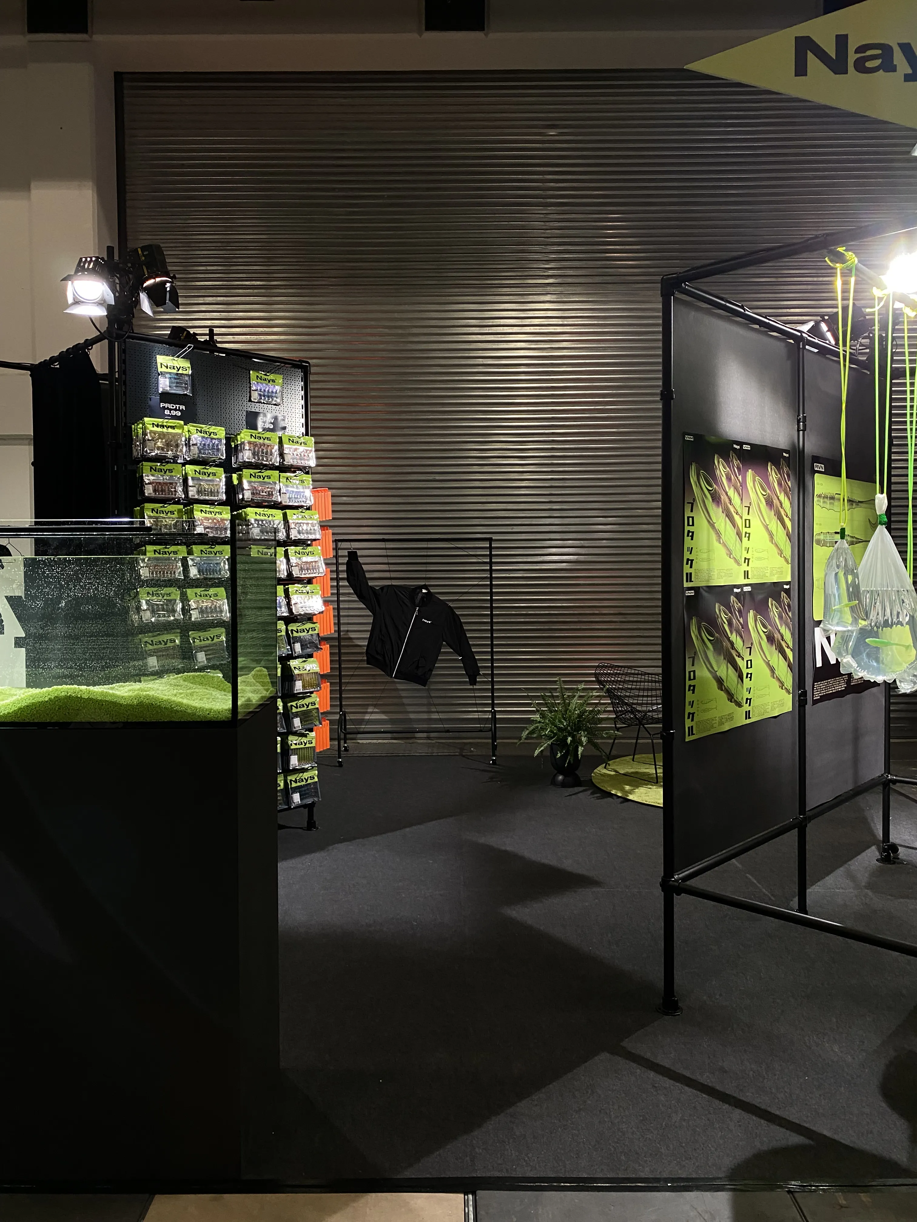 Overview of the Nays trade fair booth with aquarium, packaging tower and scaffold installation in front of a corrugated metal wall.