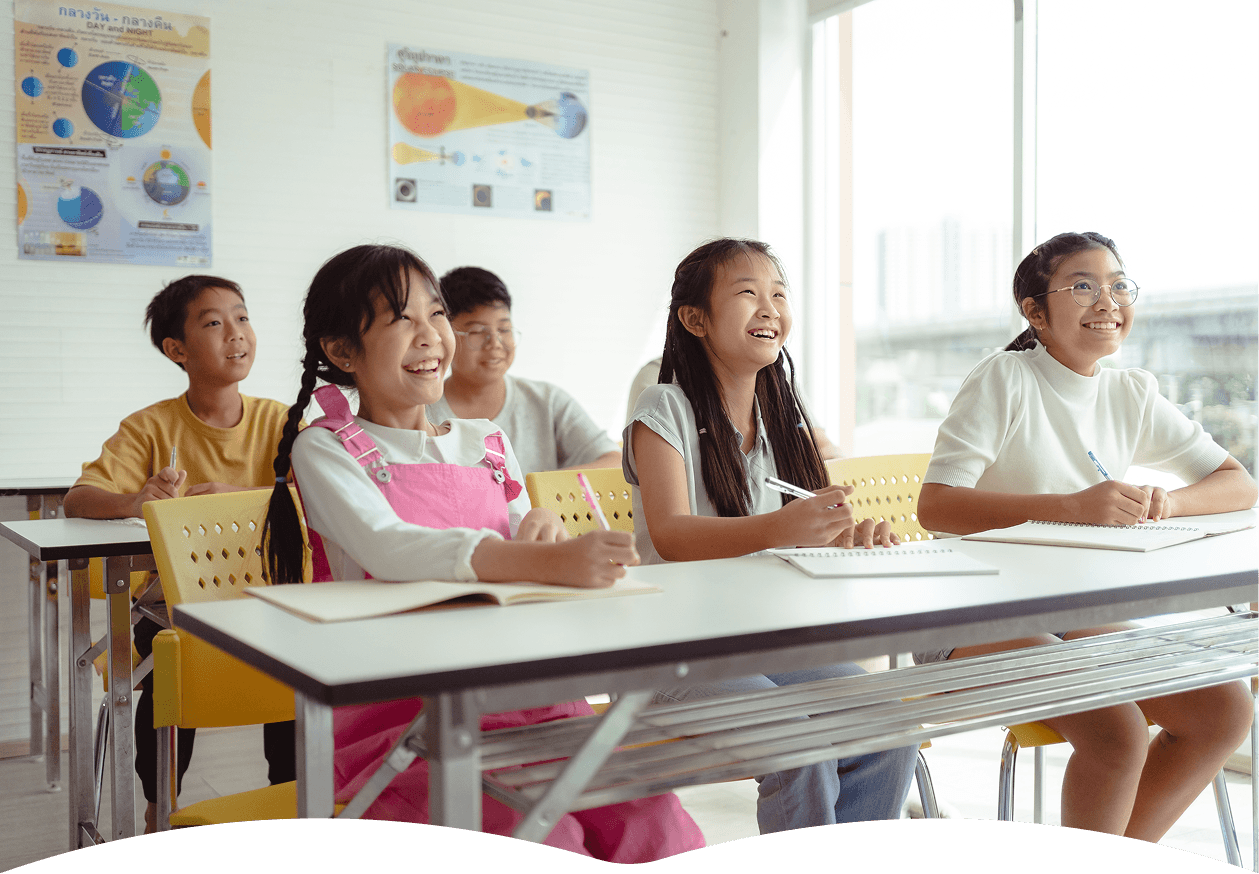 Five students, seated at desks in a bright classroom, attentively face forward while taking notes, with educational posters and large windows in the background.