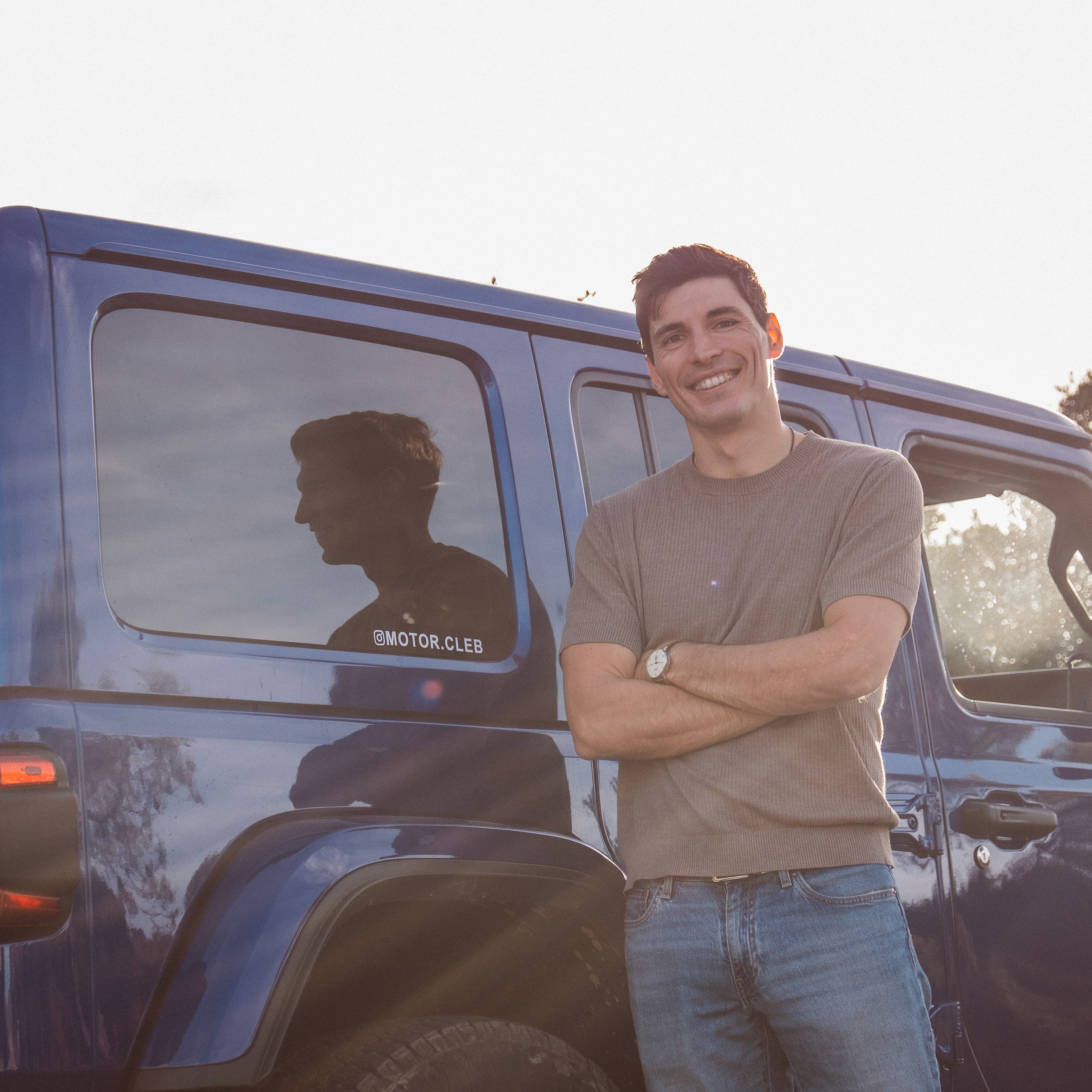 Two people in a workshop, one standing and smiling while the other leans over a car, working on it.